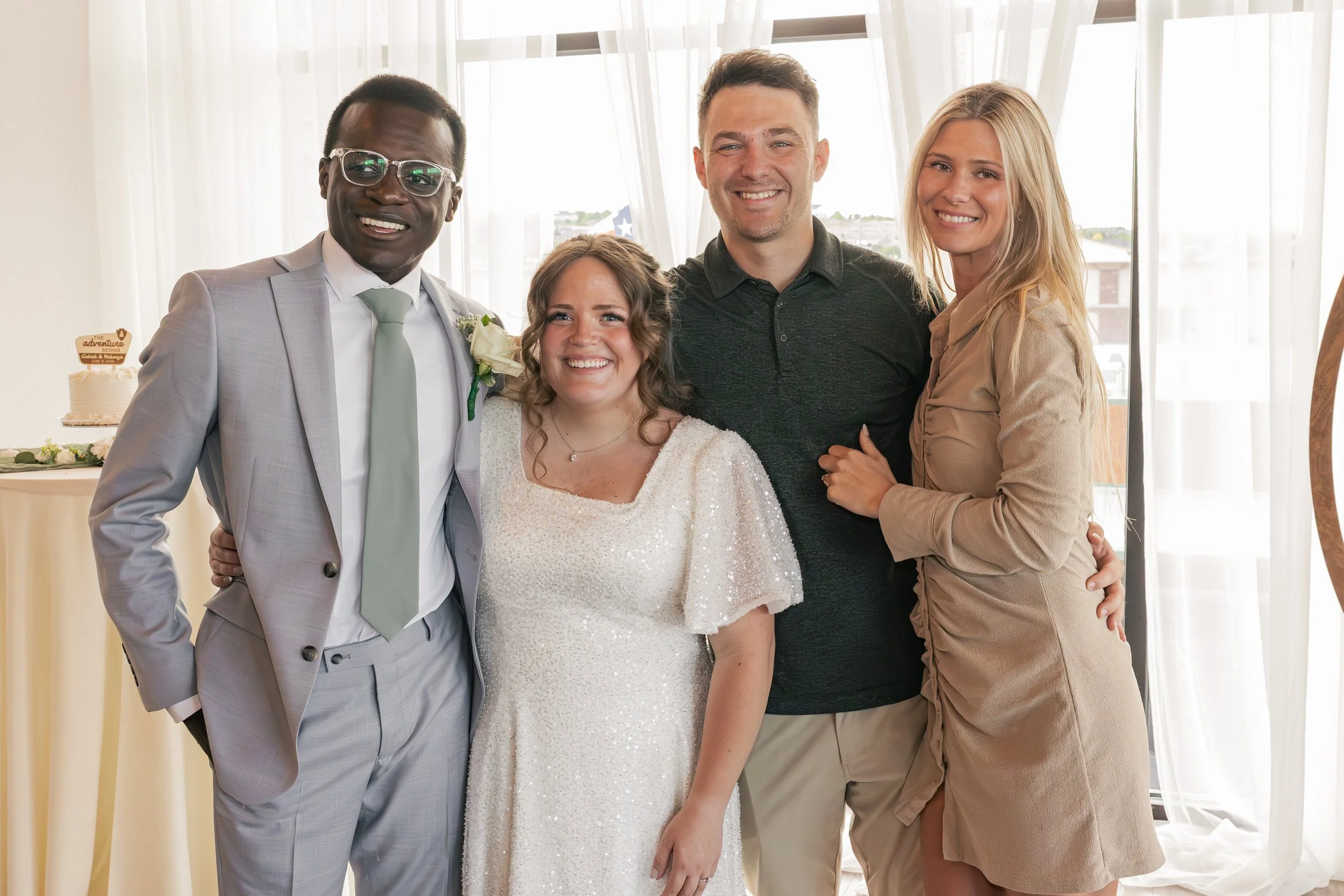 Group of five people smiling at a wedding reception, including the bride in a white dress, the groom in a light gray suit, and three guests in casual attire, standing together indoors with large windows in the background.