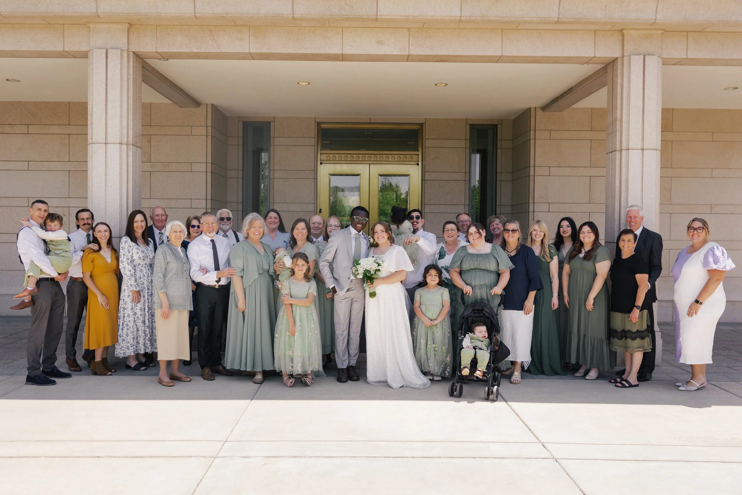 A large group of people gathered outside a building, including a bride and groom in the center, celebrating a wedding.