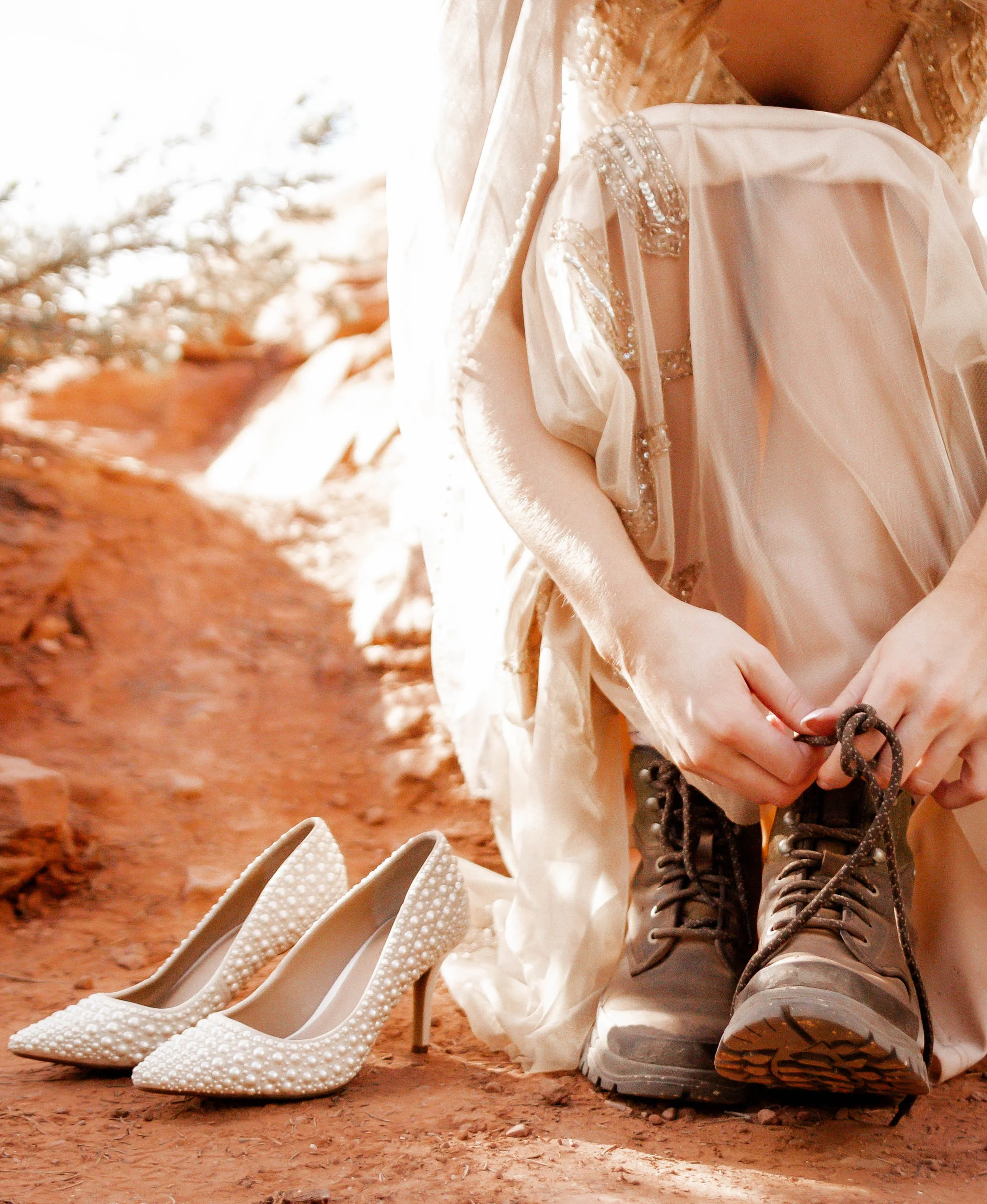 Person in a wedding dress tying the laces of hiking boots, with a pair of white high-heeled shoes with pearls placed on the ground nearby, outdoors on a reddish dirt path.