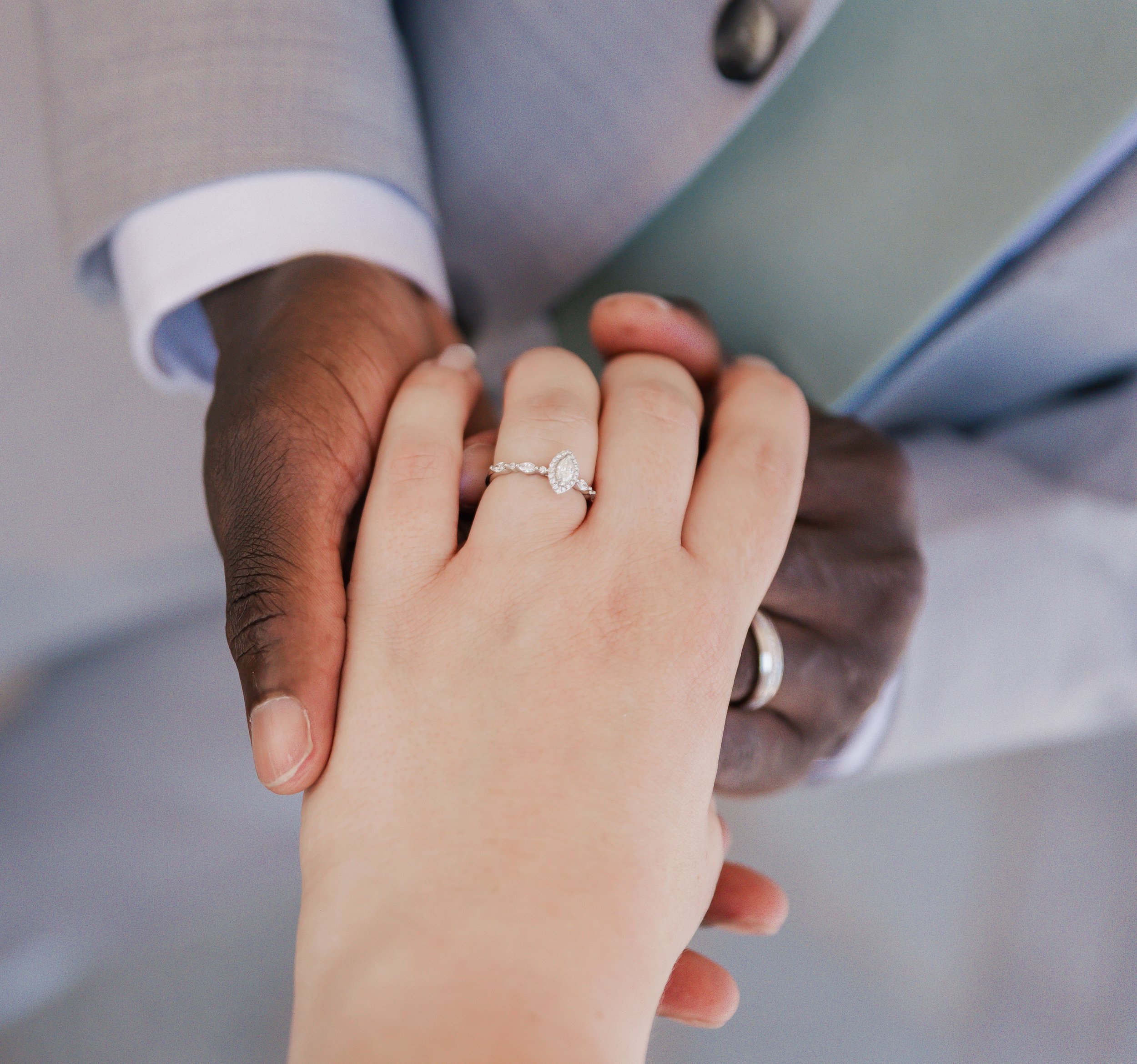 A close-up of a couple's hands holding each other. The woman's hand showcases a diamond engagement ring and a wedding band. The man's hand is darker skin and he wears a wedding ring.