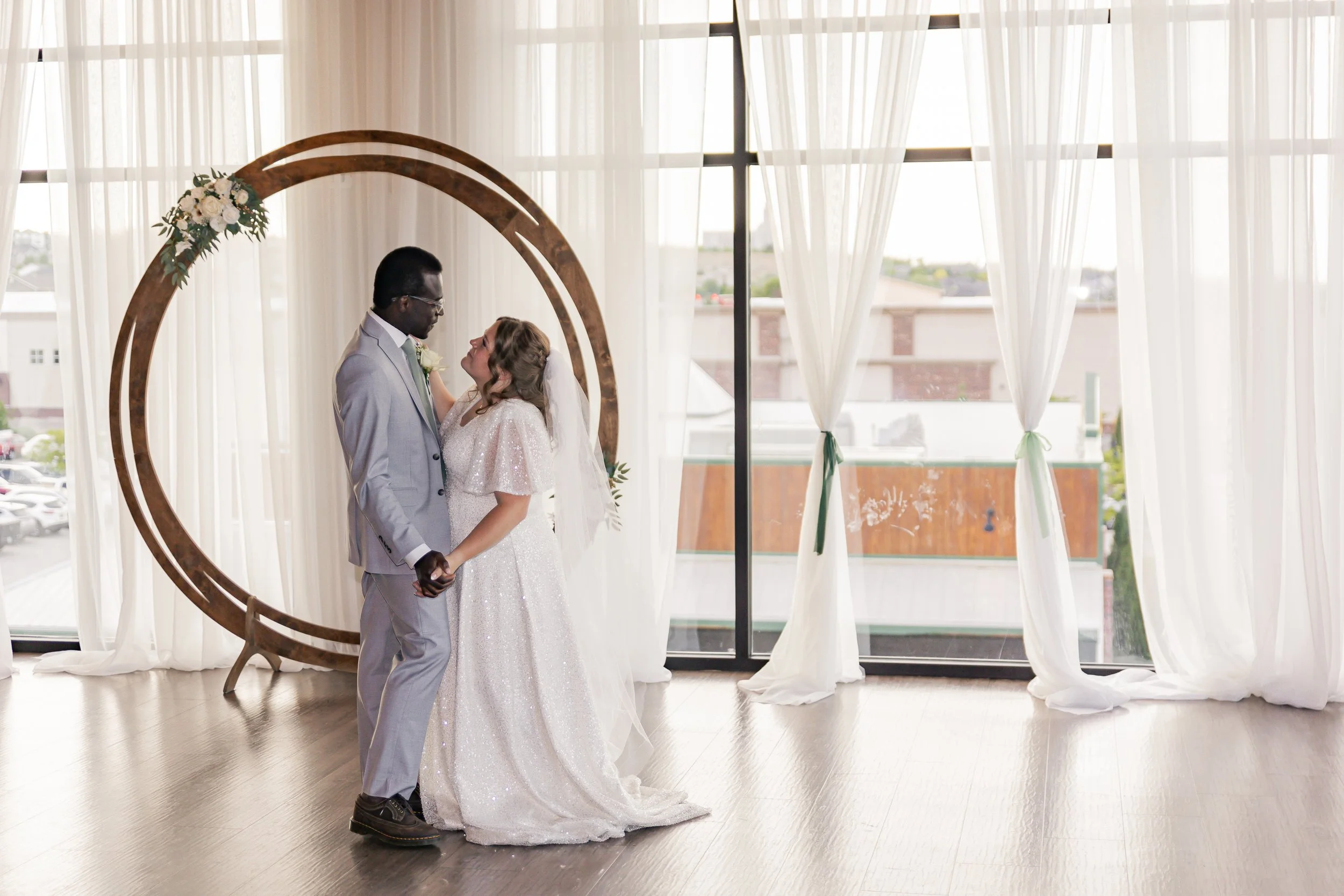 A bride and groom holding hands and looking at each other inside a wedding venue with large windows and sheer white curtains, standing in front of a circular wooden arch decorated with flowers.