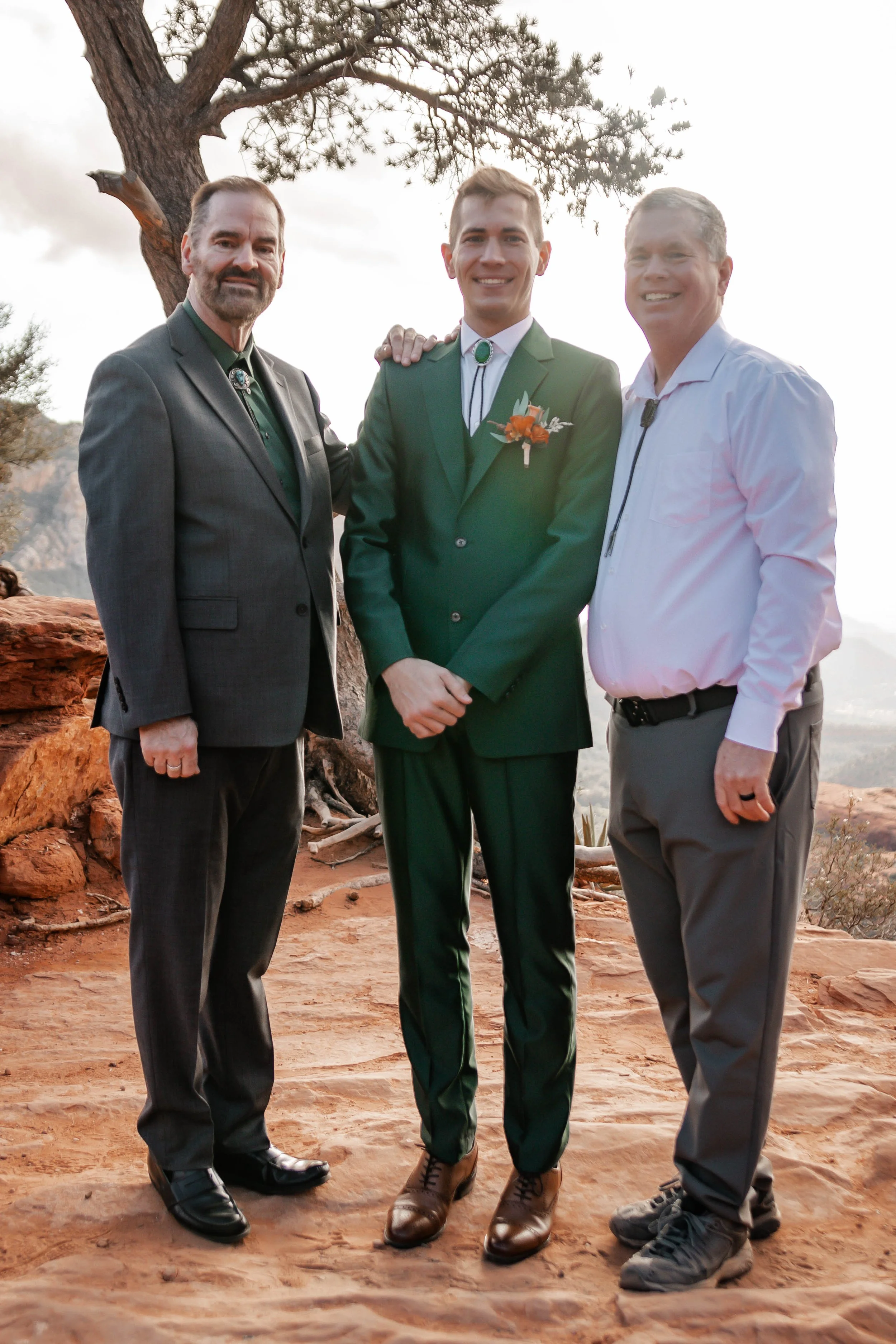 Three men in formal attire standing outdoors on reddish rock with a tree and cloudy sky in the background. The man in the middle is wearing a dark green suit with a boutonniere, while the two men on either side are wearing lighter-colored shirts, and