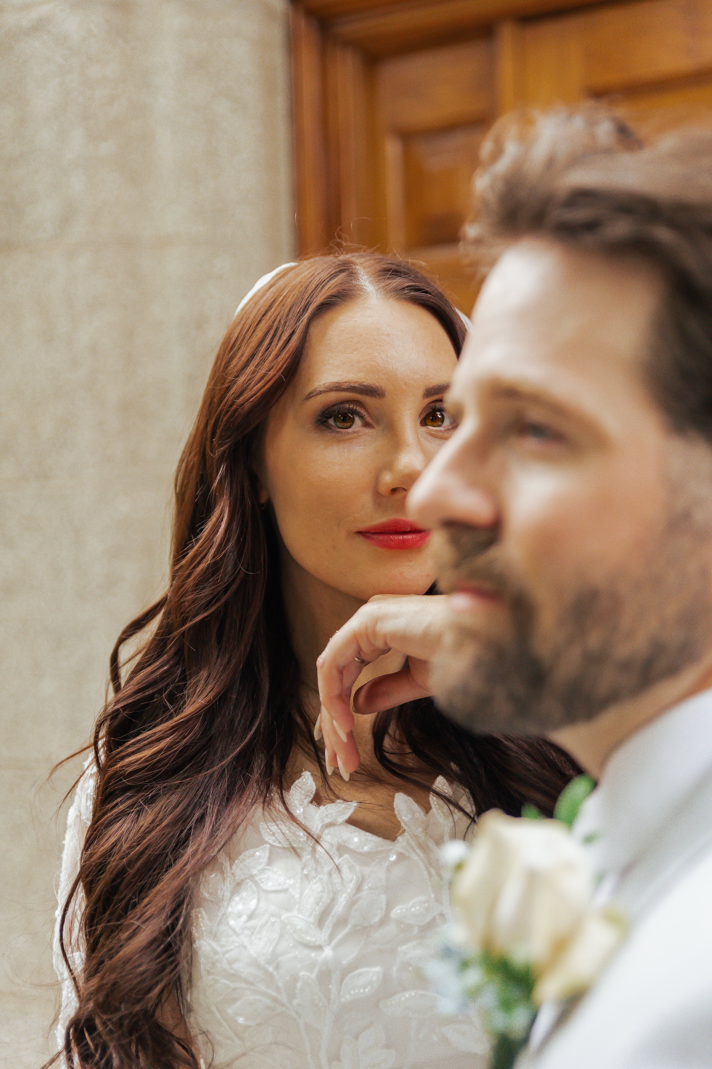 A woman with long brown hair and red lipstick looks directly at the camera, while a man with a beard and mustache, dressed in a tuxedo with a boutonniere, is in the foreground and slightly blurred.