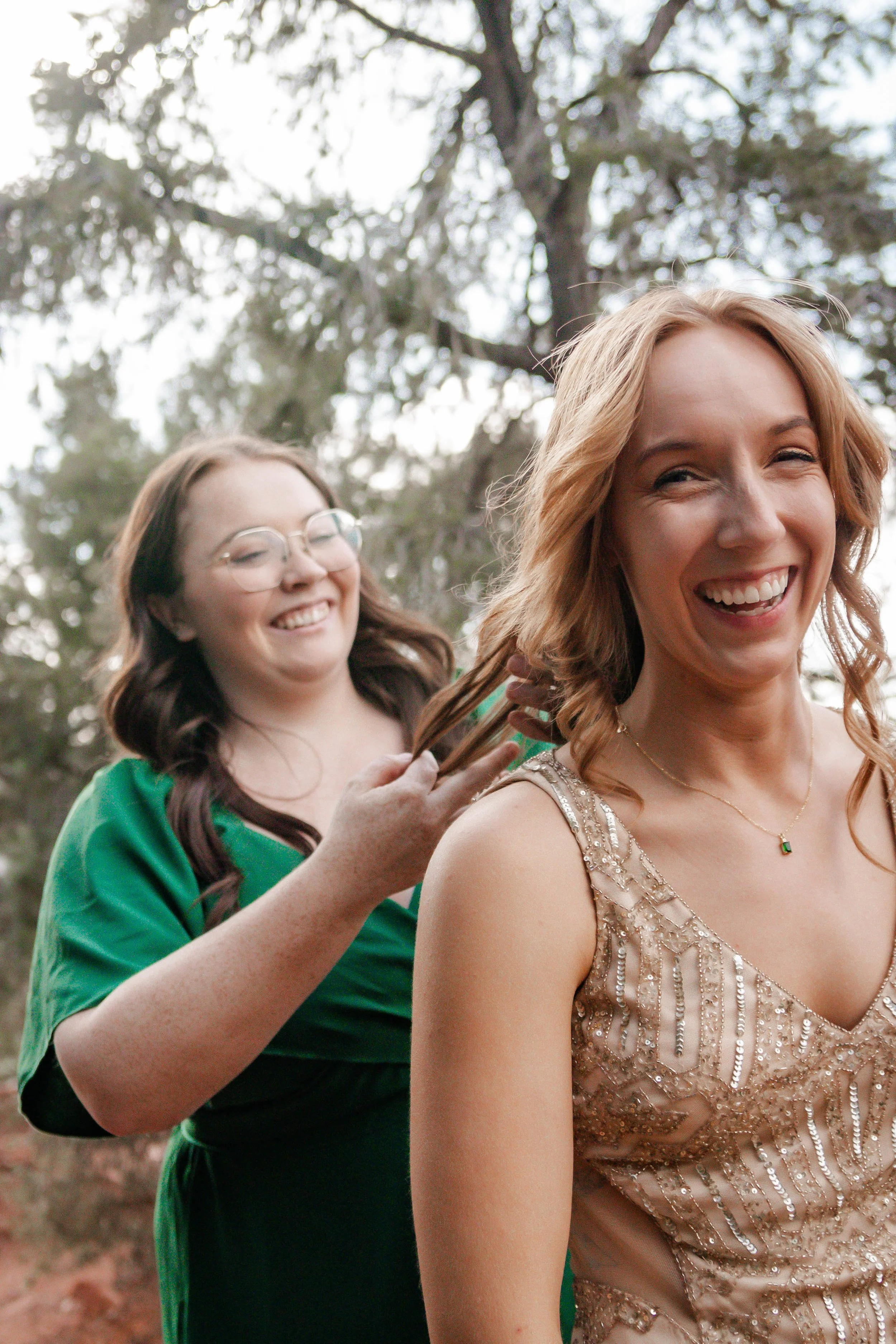 Two women outdoors, one with red hair in a sparkly dress smiling, the other with brown hair and glasses in a green dress, touching her hair, with trees in the background.