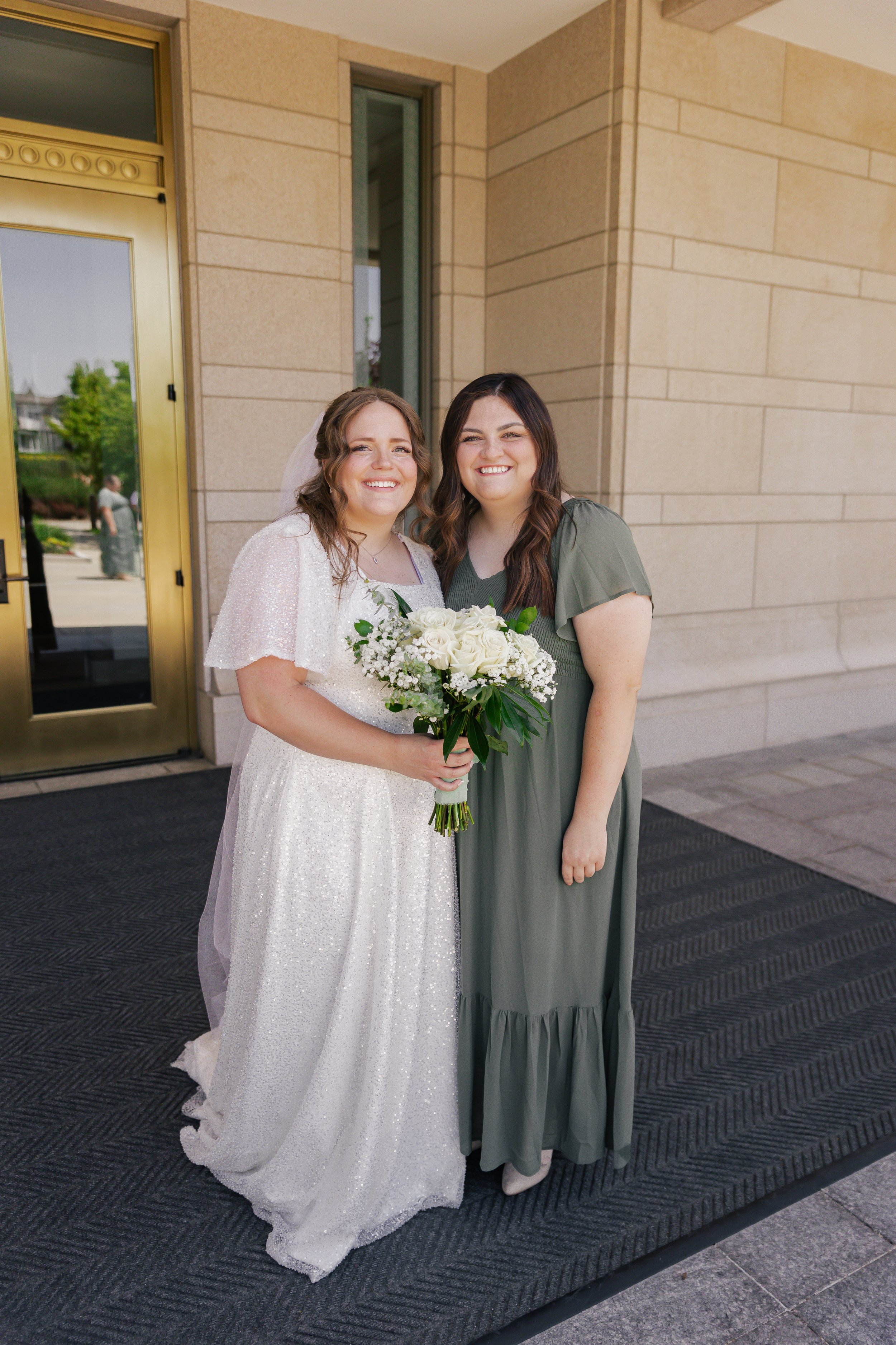 A bride in a white wedding dress holding a bouquet of white roses and greenery, standing next to a woman in a long green dress outside a building with beige stone walls and a gold door.