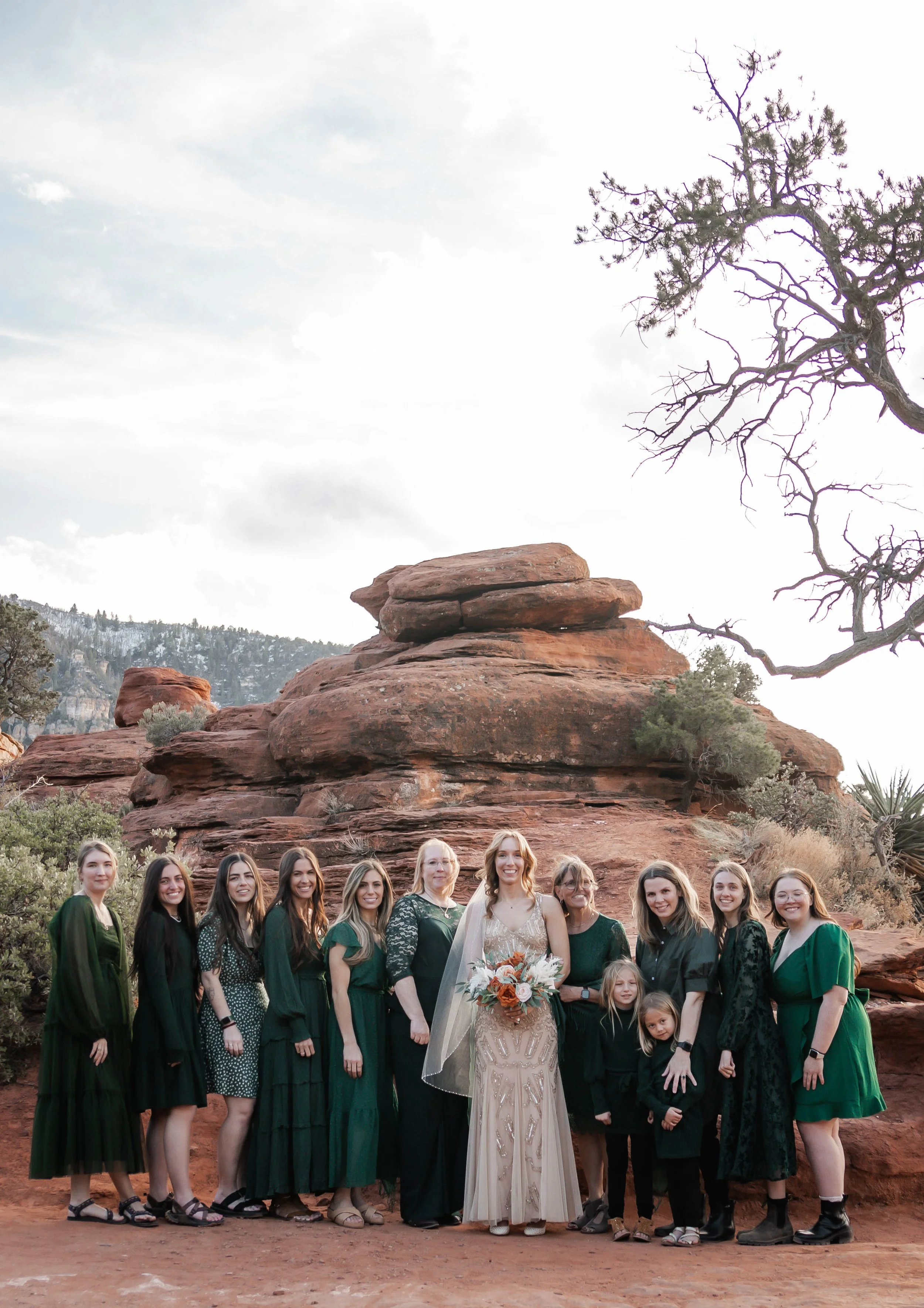 Group of women in dark and green dresses, including a bride holding a bouquet, standing in front of red rock formations in a desert landscape.