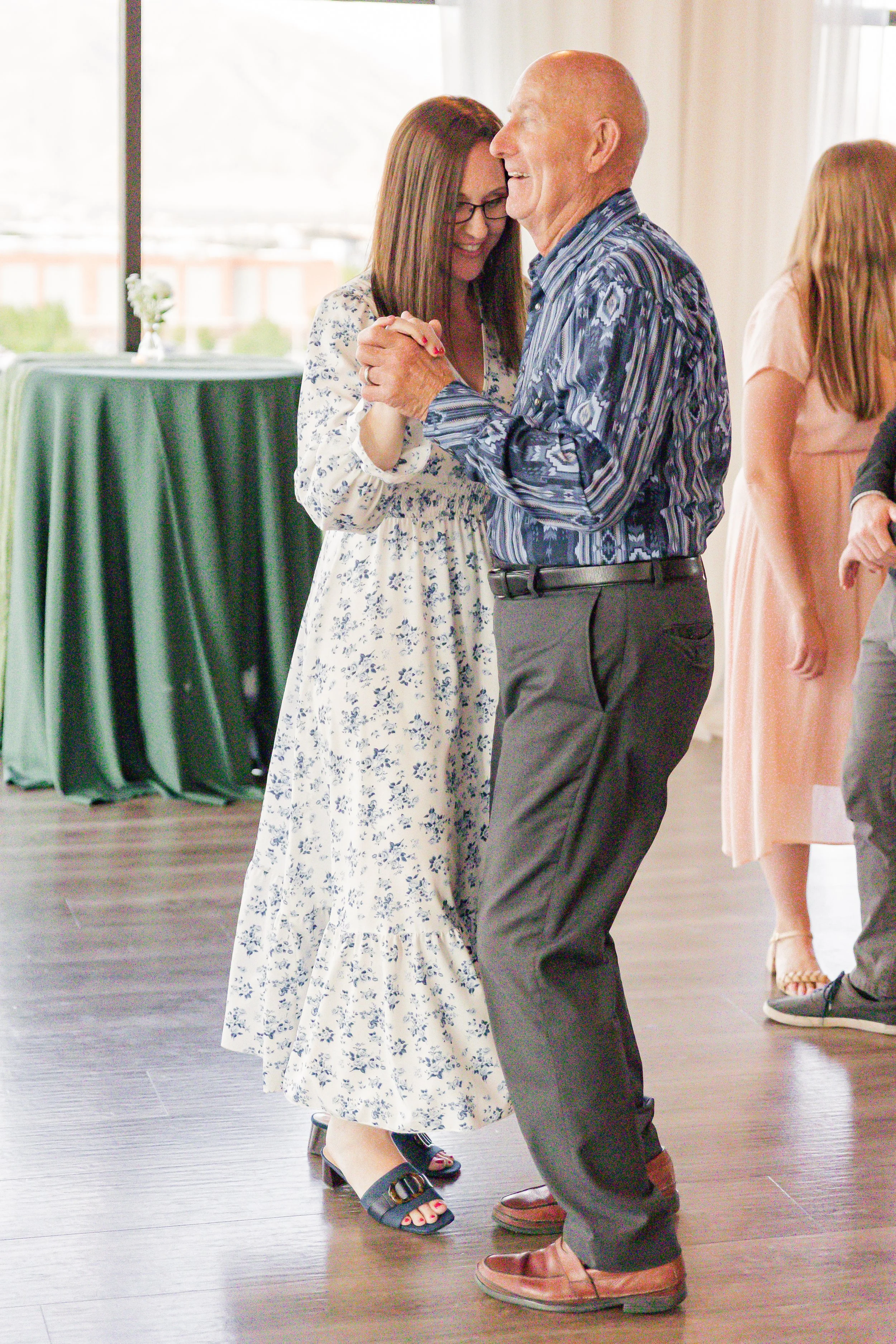 A man and woman are dancing together inside a bright room, holding hands and smiling. The woman wears a floral dress and glasses, and the man wears a patterned shirt and dark pants.