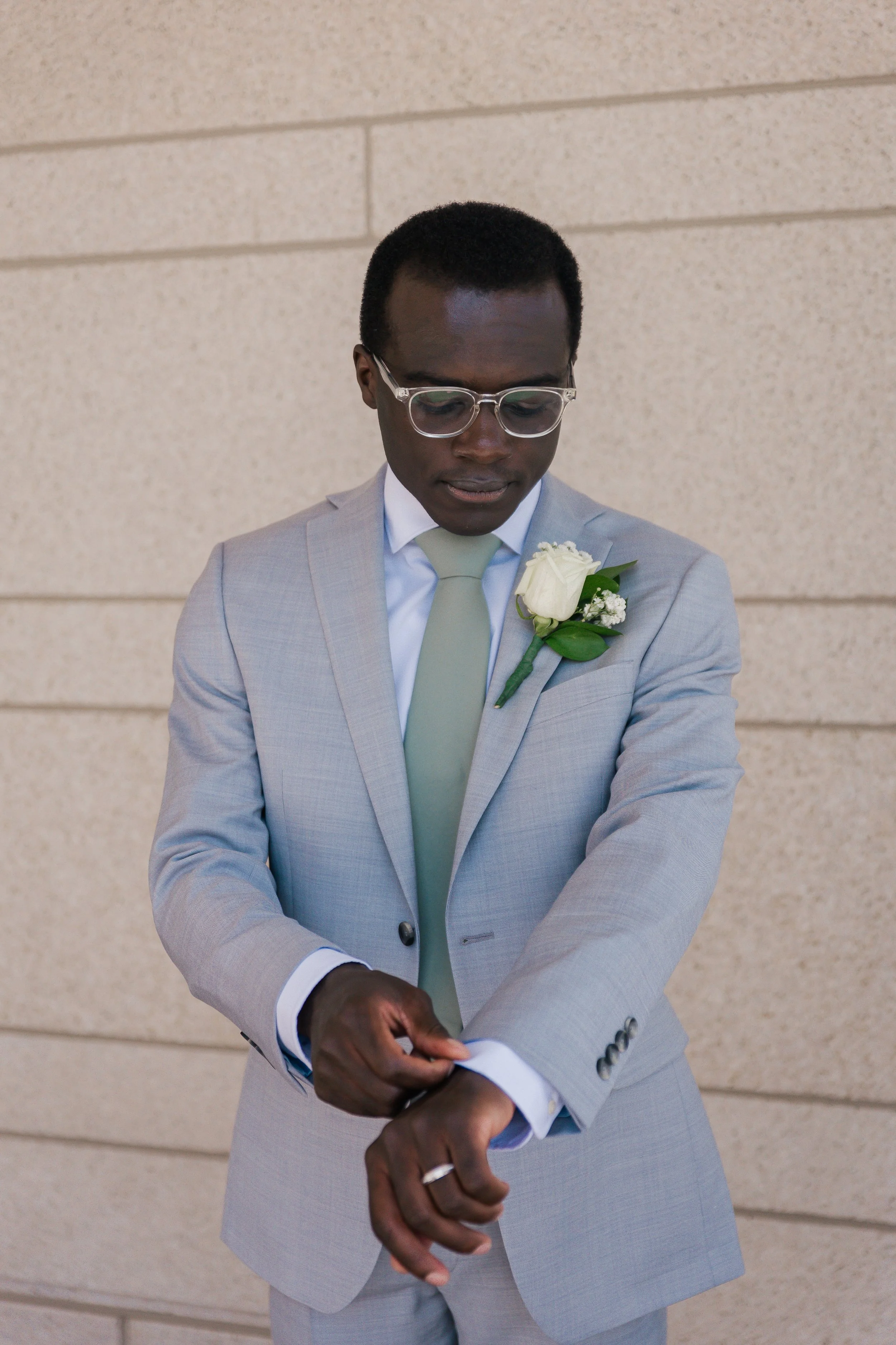 A man in a light gray suit with a white shirt and light green tie, wearing glasses, is adjusting his sleeve. He has a white boutonniere pinned to his suit lapel and is standing against a beige brick wall.