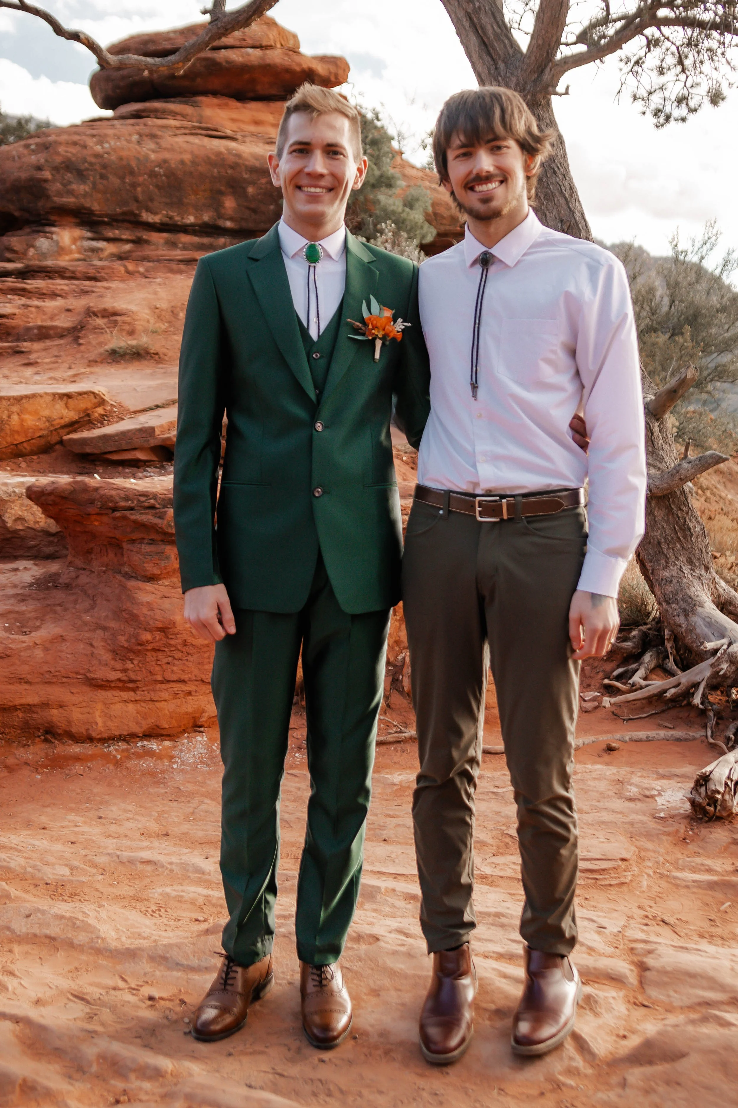 Two men standing on red rocky terrain, smiling at the camera. One is wearing a green suit with a boutonniere, and the other a white shirt and khaki pants. Behind them are trees and a cloudy sky.
