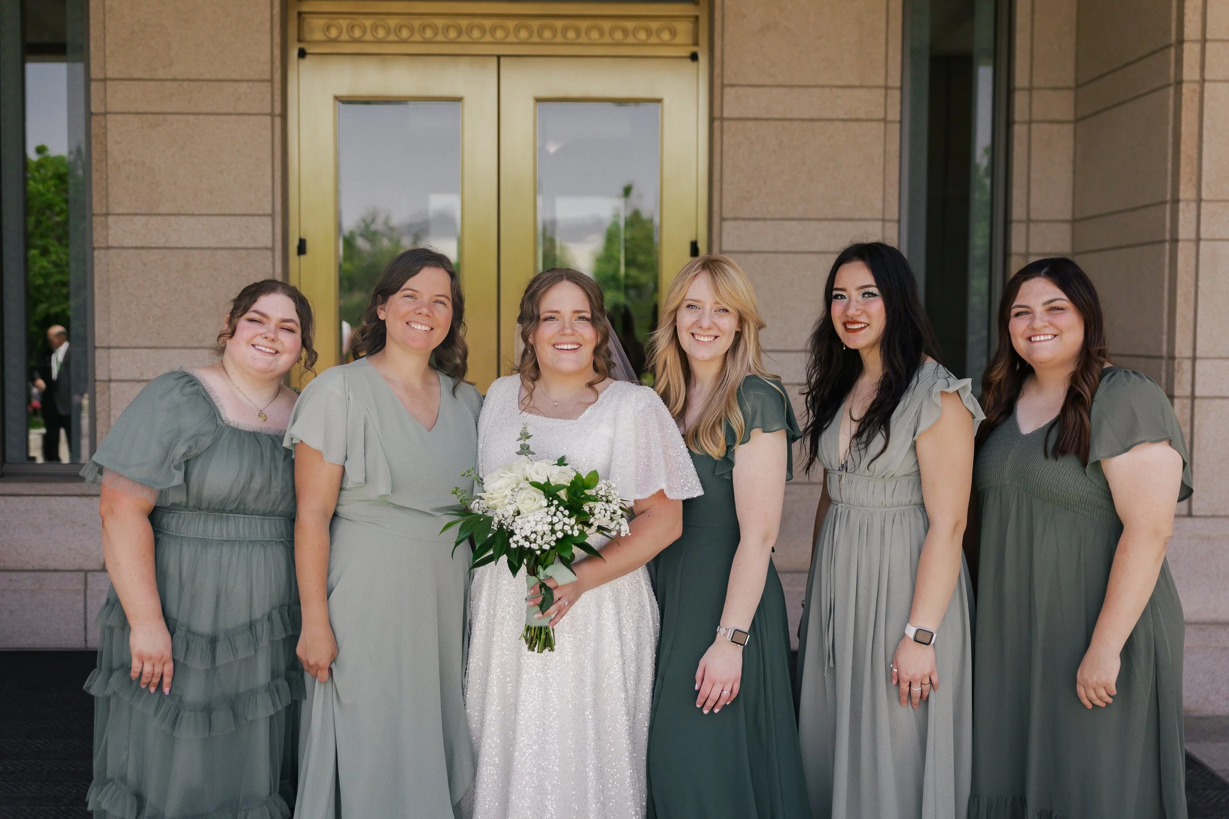 Bride in white dress holding bouquet with five bridesmaids in green dresses standing outside in front of a building with large gold double doors.