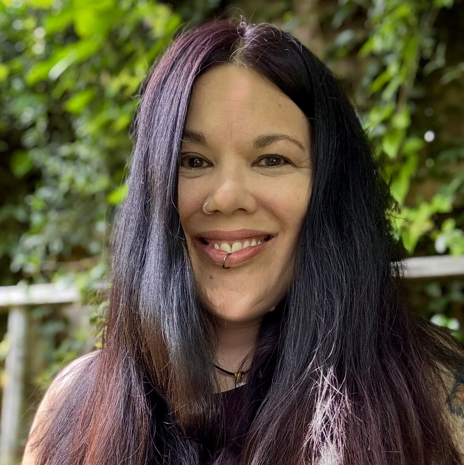 A woman with long black hair featuring purple and pink highlights, smiling outdoors with a background of greenery.