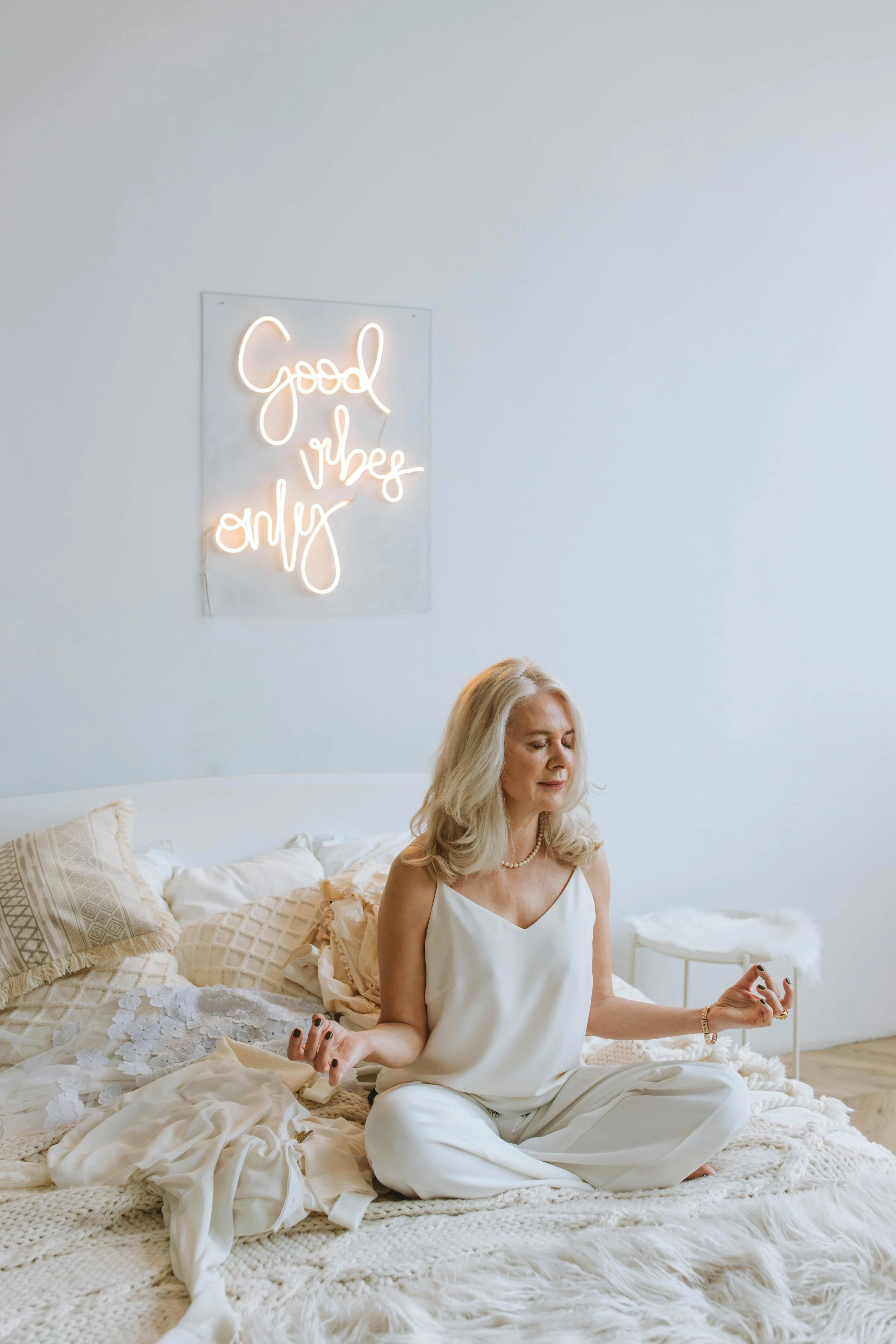 Woman sitting cross-legged on a cozy bed, meditating peacefully under a neon sign that says “Good vibes only.” The bright, calming setting evokes mindfulness, self-care, and emotional wellness.