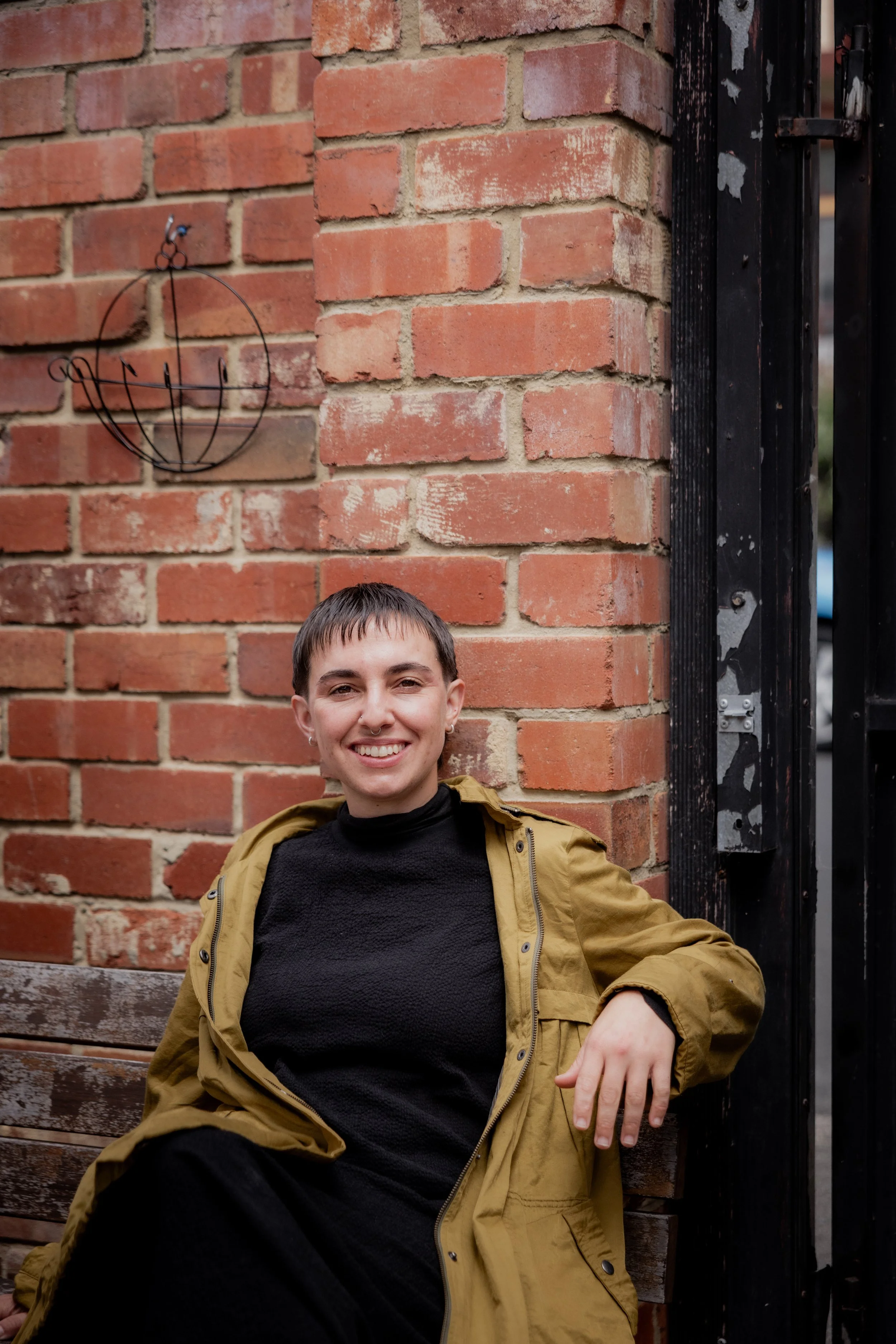 A smiling person sitting on a wooden bench against a brick wall, wearing a black top and a tan jacket, with metal artwork on the wall behind them.