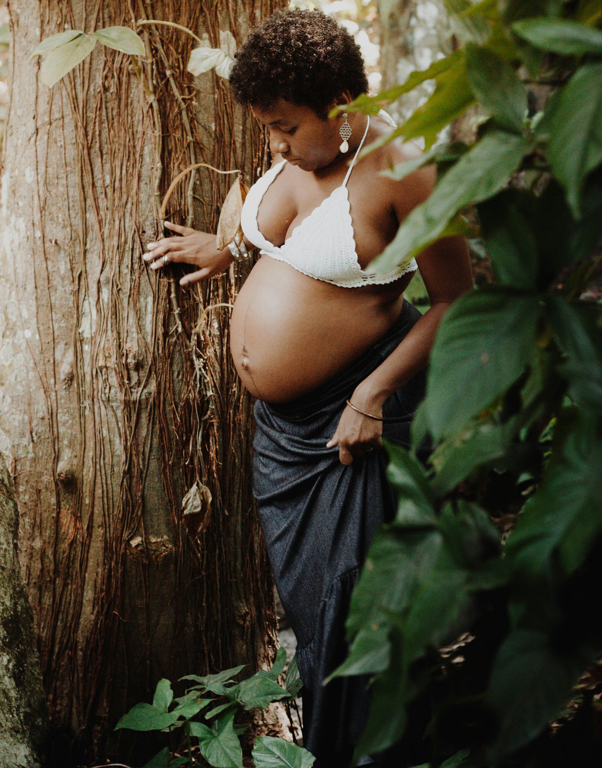 A pregnant woman in a white crochet top and dark pants standing next to a tree in a lush green environment.