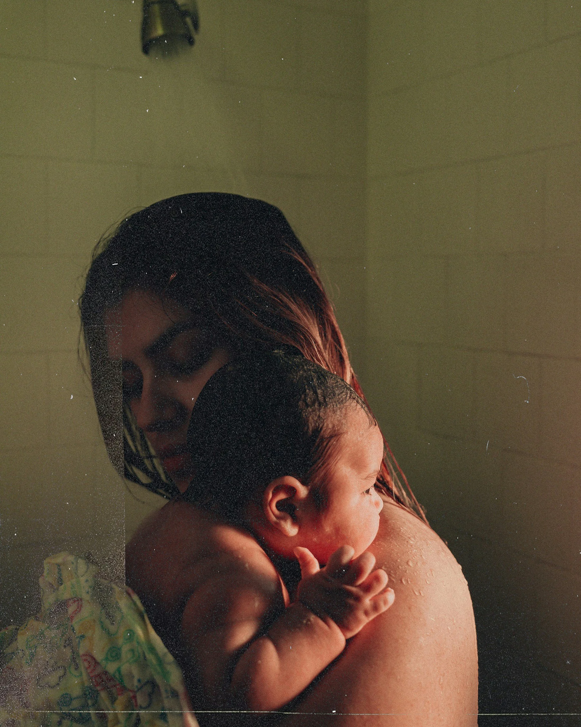 A woman holding a baby during a bath, with her face partly visible, as she cradles the baby close to her shoulder in a shower setting.
