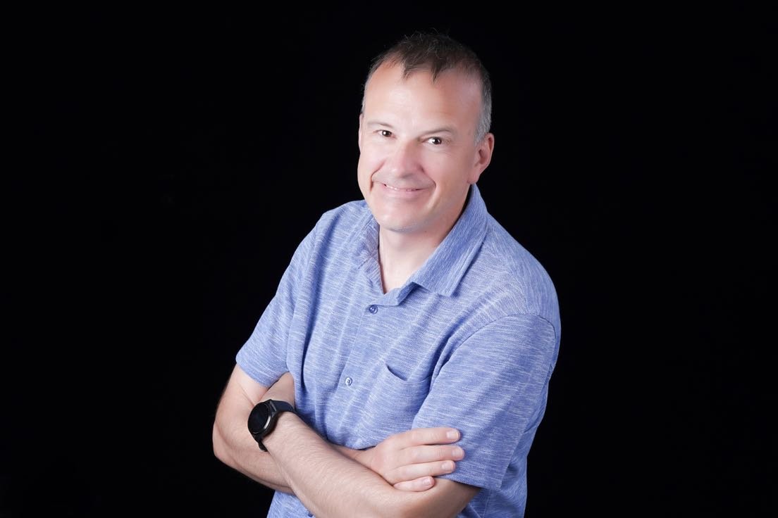 A man with short brown hair, wearing a blue polo shirt and a black watch, standing with arms crossed and smiling against a black background.