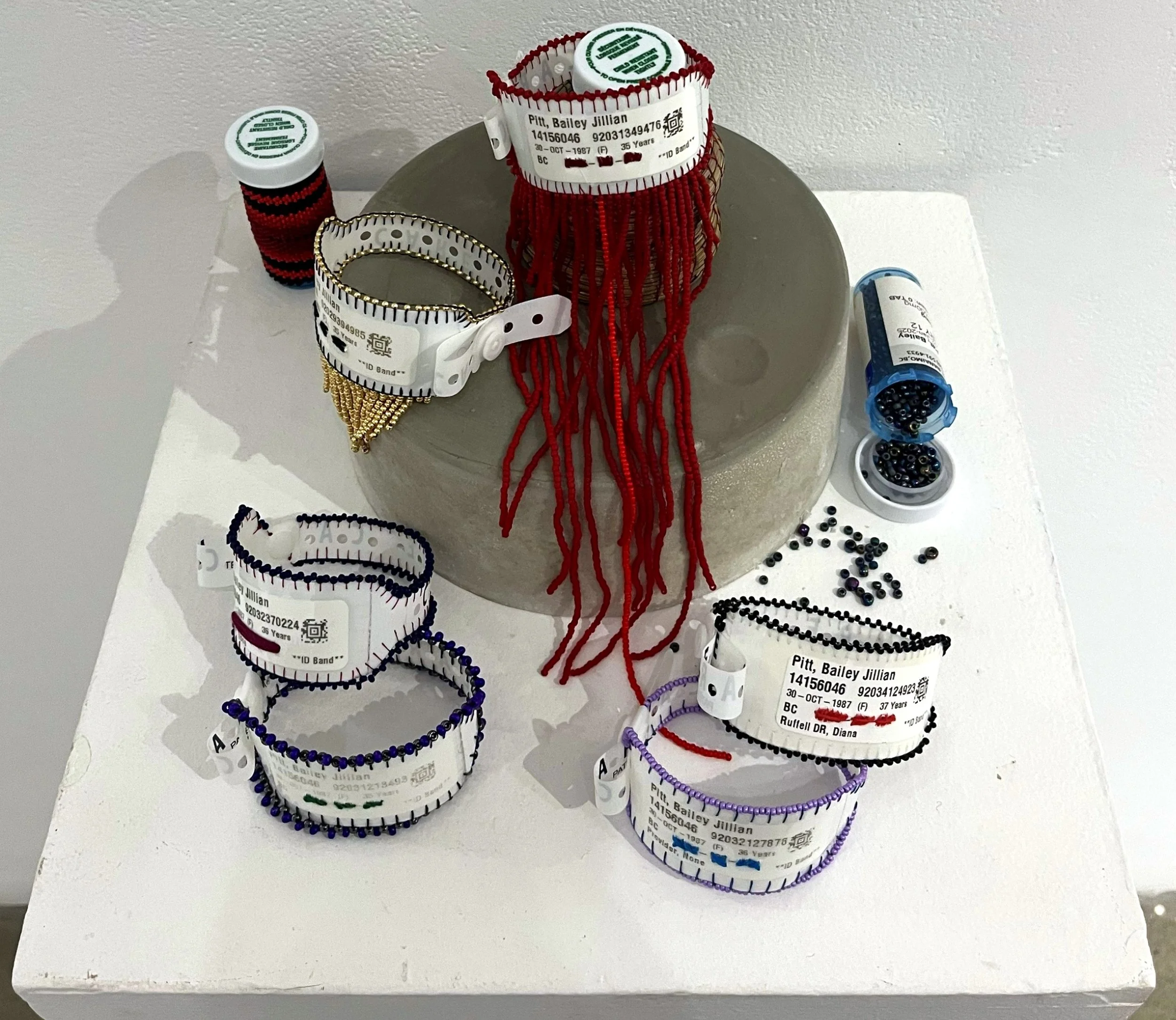 A display of colorful beaded bracelets and a container of small black beads on a white surface. The bracelets have labels resembling hospital bracelets with names, dates, and barcodes.