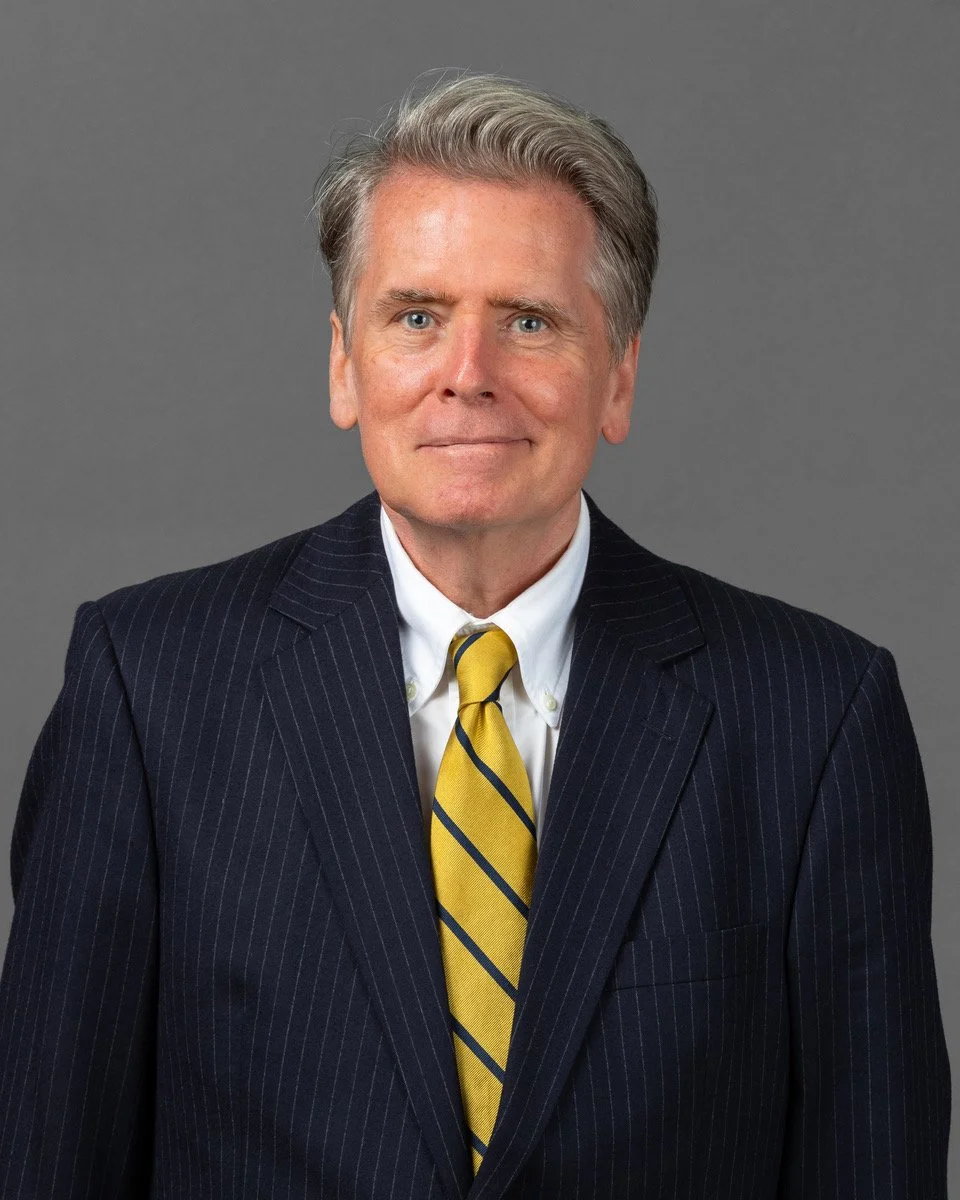 A man with gray hair in a suit and tie standing in front of bookshelves filled with books and a small red decorative object.