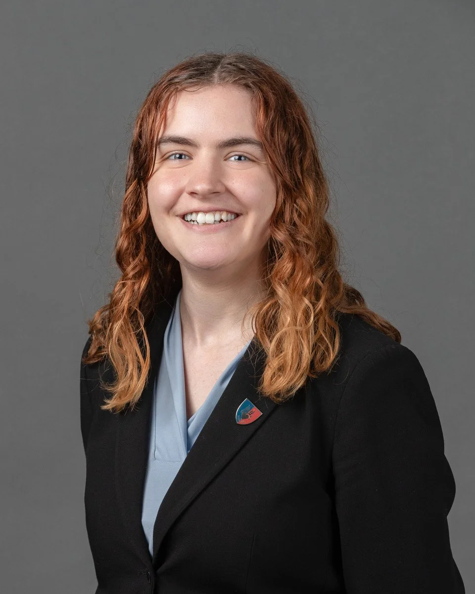 Young woman with curly red hair smiling in front of a dark blue backdrop with white text and logo, wearing an orange ribbed sweater.
