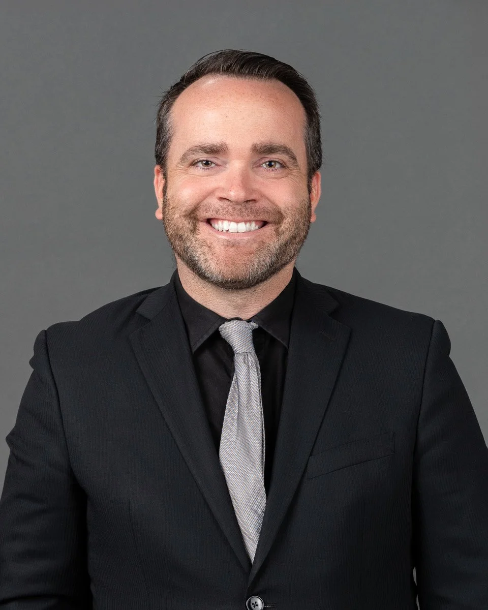A man in a beige blazer and a pastel checkered shirt smiling in front of a blue backdrop with logos for The Chesterton Schools Network.