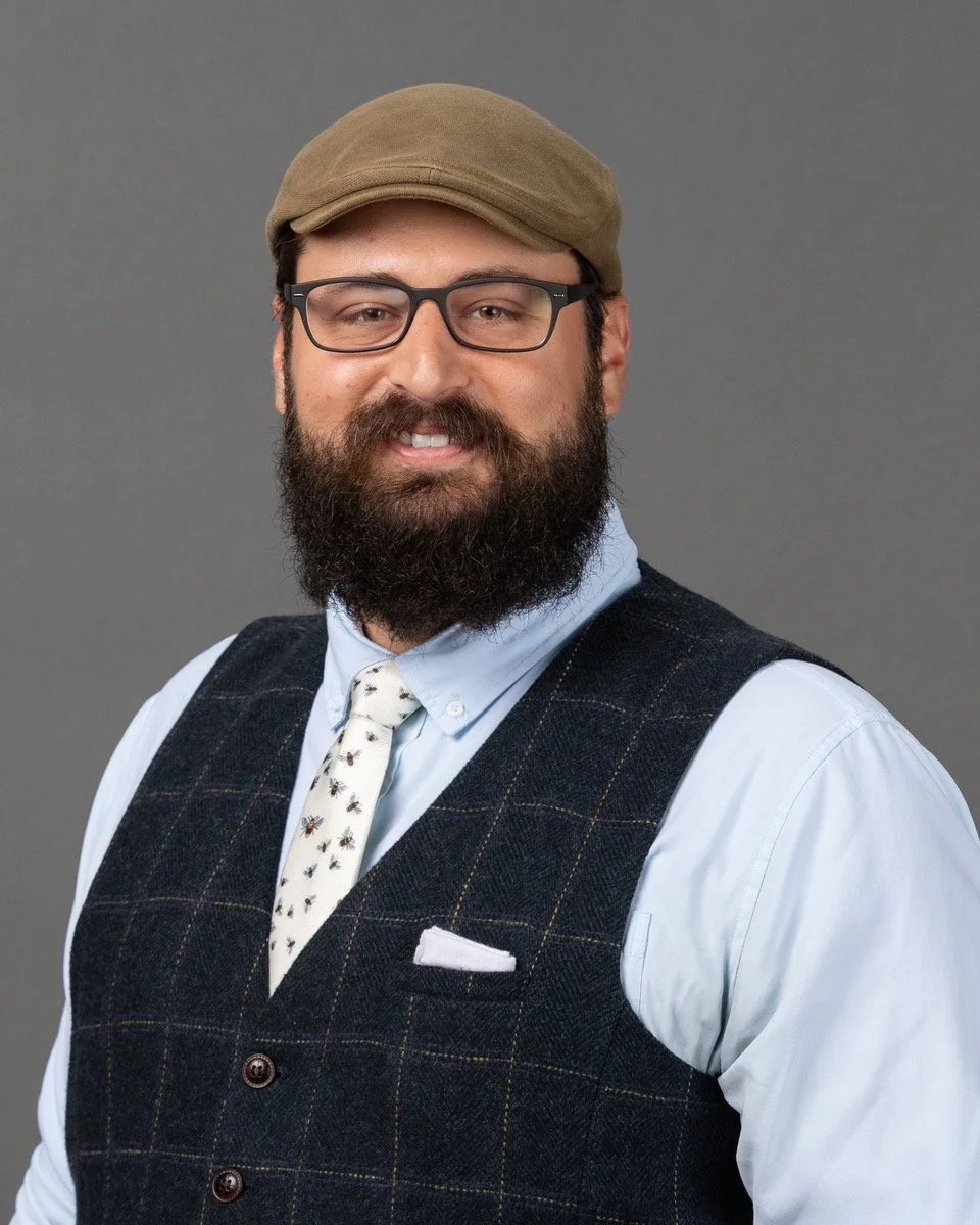 A man with a full beard and mustache, wearing a plaid shirt, standing in front of a blue backdrop with the logo and text of The Chesterton Schools Network, Joyfully Catholic, Classical High Schools.