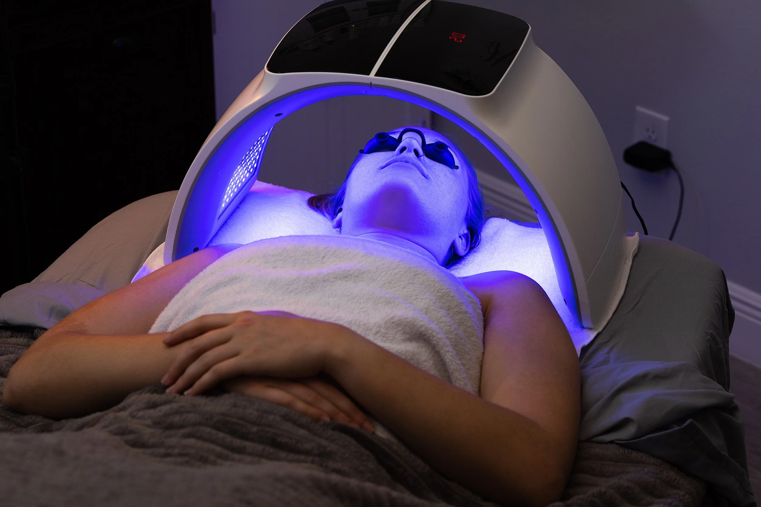 Person lying on a bed undergoing LED light therapy with a blue light mask connected to a machine, covered with a towel, in a dimly lit room.