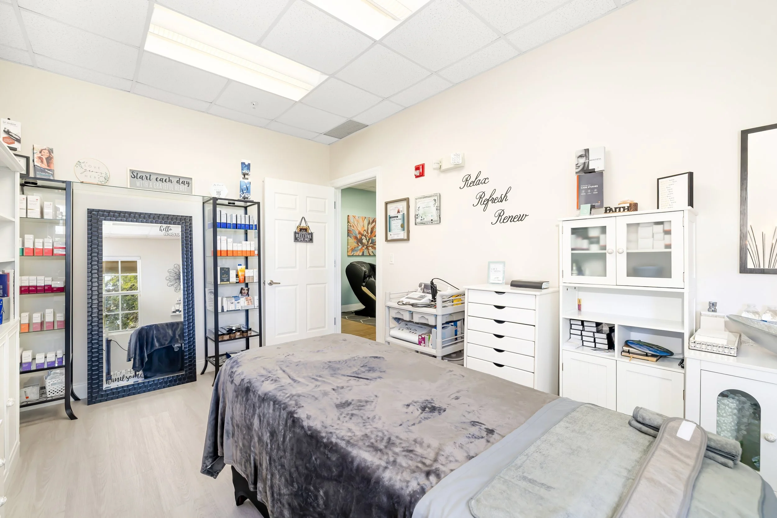 Interior of a wellness clinic or spa room with a massage table, shelves of skincare or medication bottles, a large mirror, framed certificates, and inspirational wall quotes. Bright lighting and calming decor. Bradenton Spa