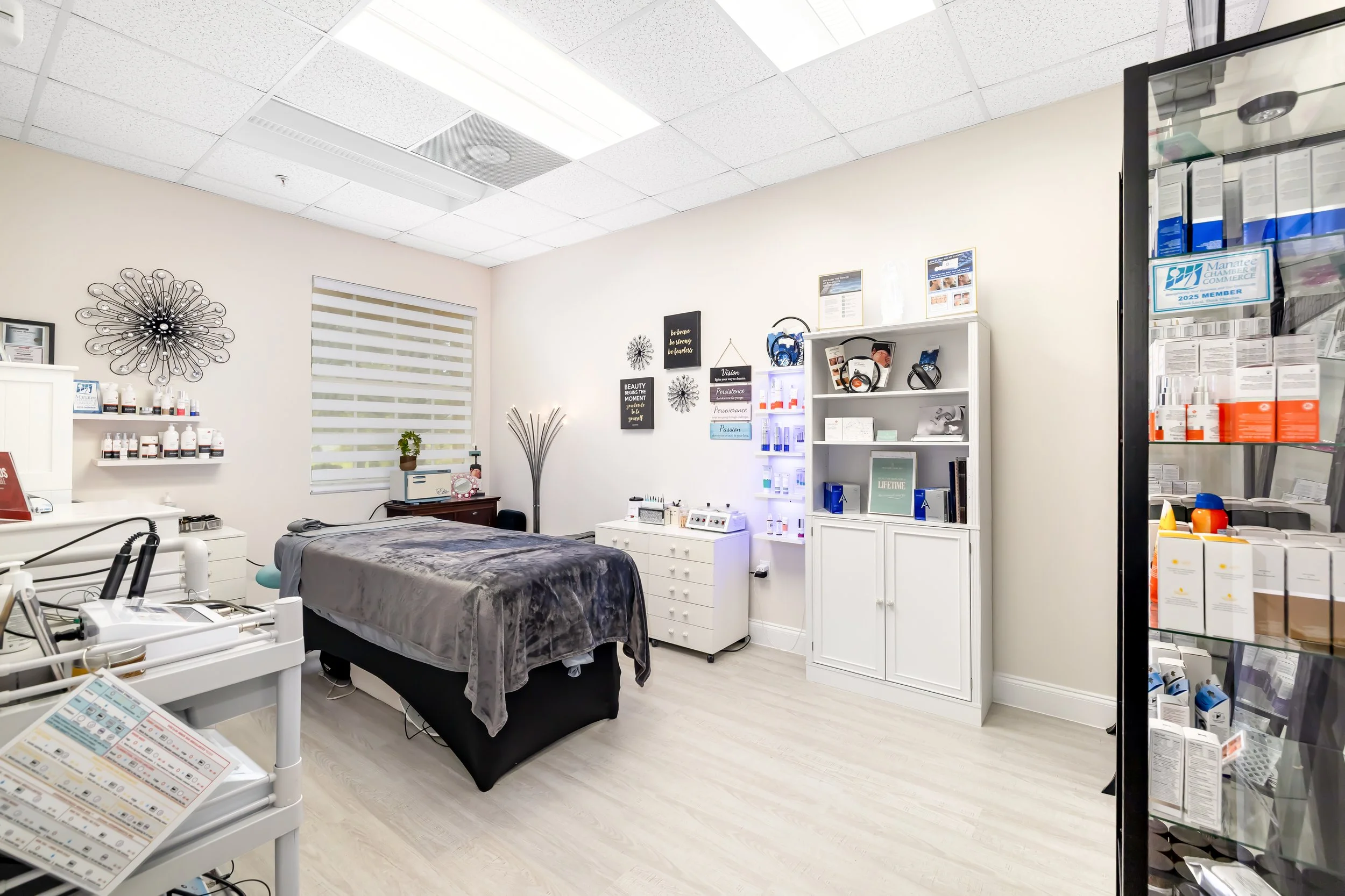 A clean, well-lit massage or treatment room with a massage bed covered in a gray blanket, surrounded by shelves and cabinets with skincare products in a health or spa clinic. Bradenton spa