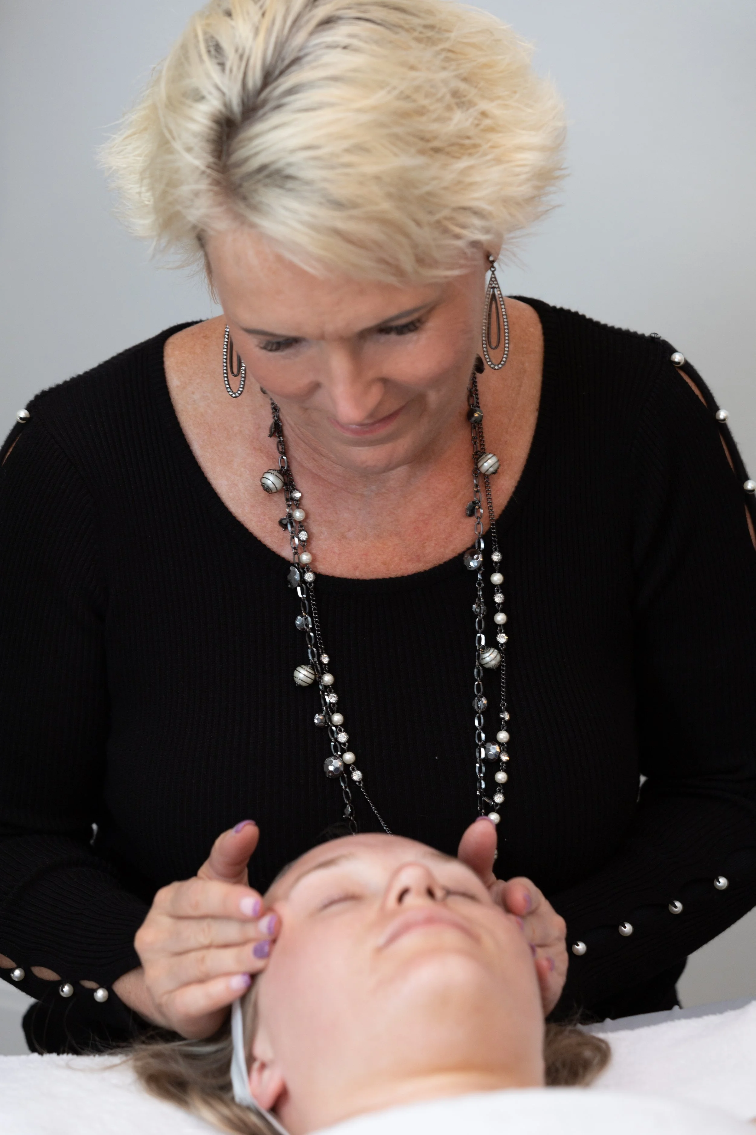 A woman with short, blonde hair giving a facial massage to a woman lying on a table with her eyes closed.
