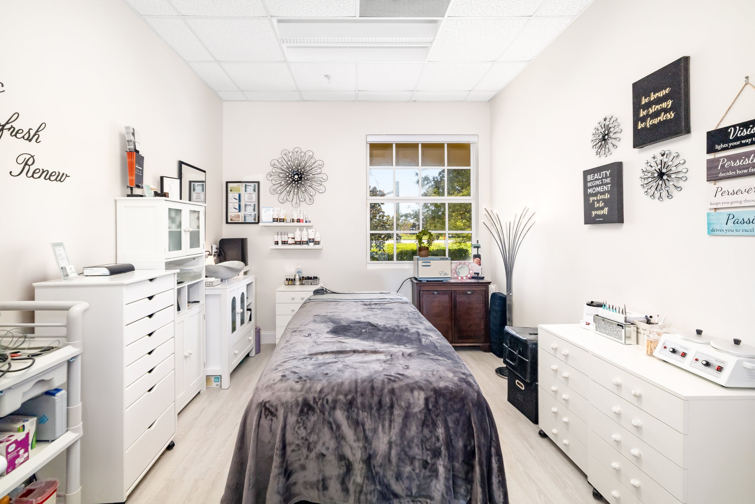 Spa or massage room with a bed covered in gray blanket, white furniture, wall art with inspirational quotes, and a window showing greenery outside.Bradenton Spa