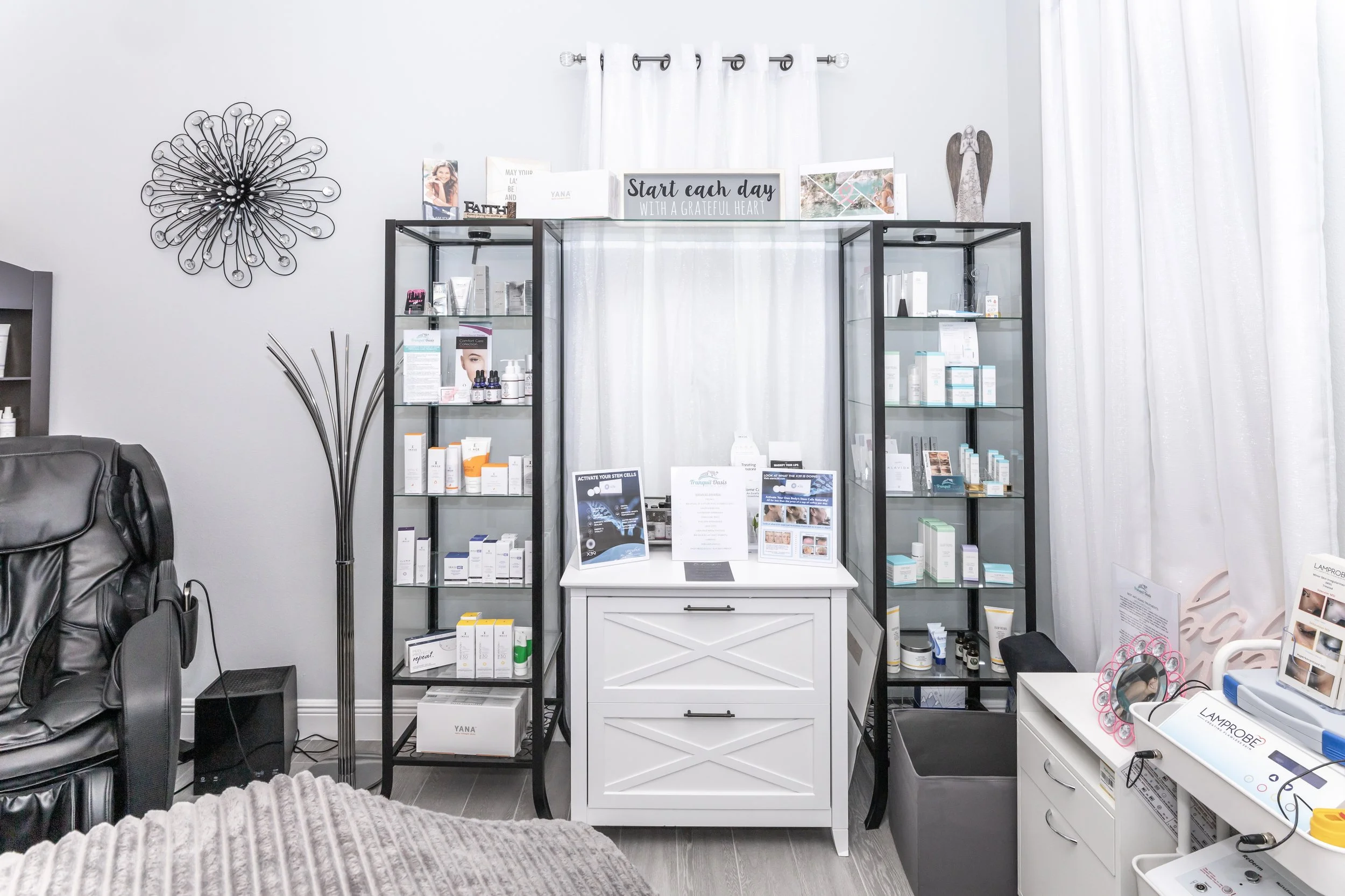 Interior view of a skincare or beauty treatment room with black display shelves filled with skincare products, a white cabinet in the center, decorative wall art including a wire flower sculpture, a white curtain, and promotional signs and brochures.