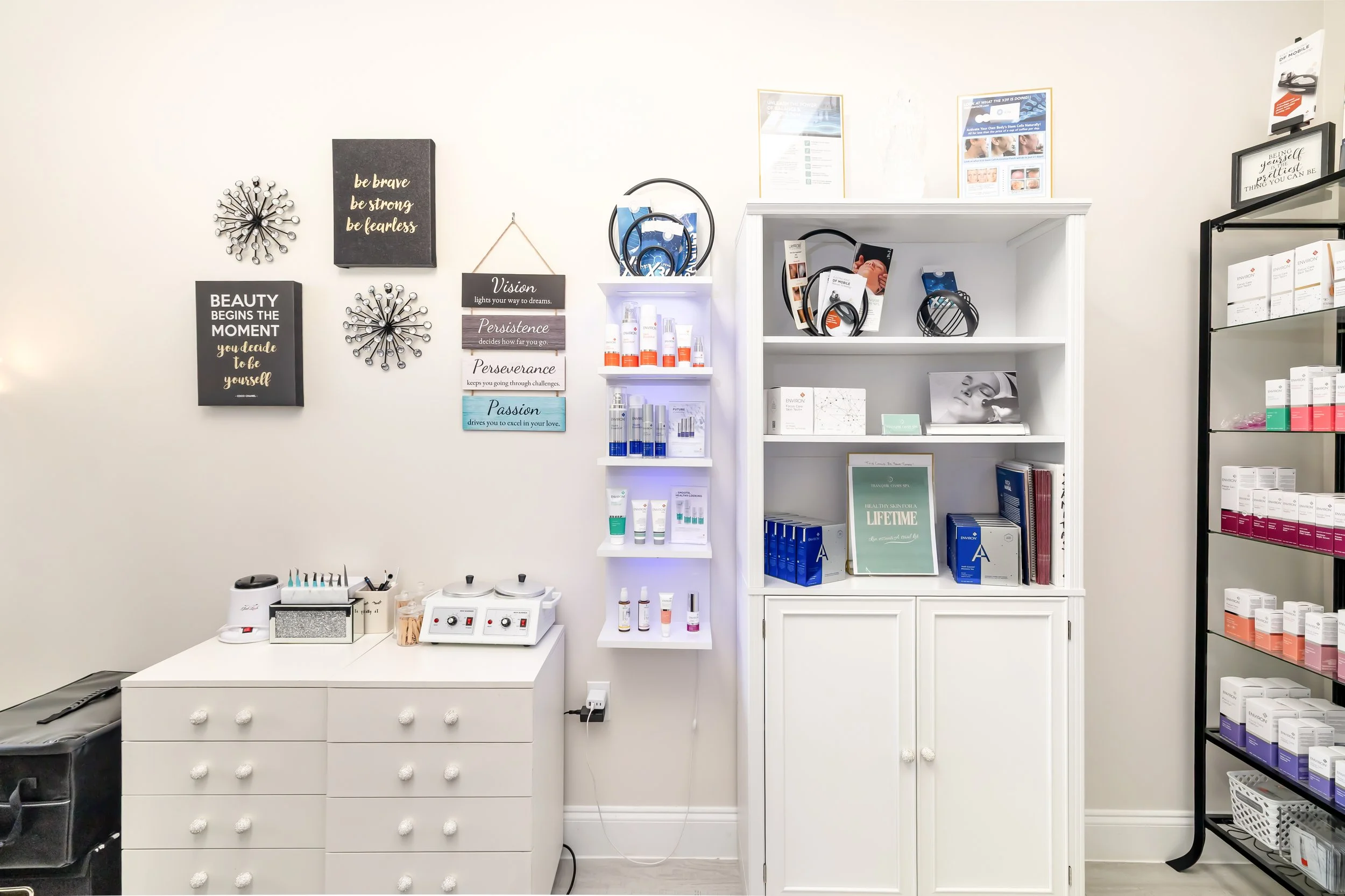 A retail or therapy room with white walls and shelves, displaying skincare or cosmetic products, motivational quotes on the wall, and various beauty tools on a white cabinet. Bradenton Spa