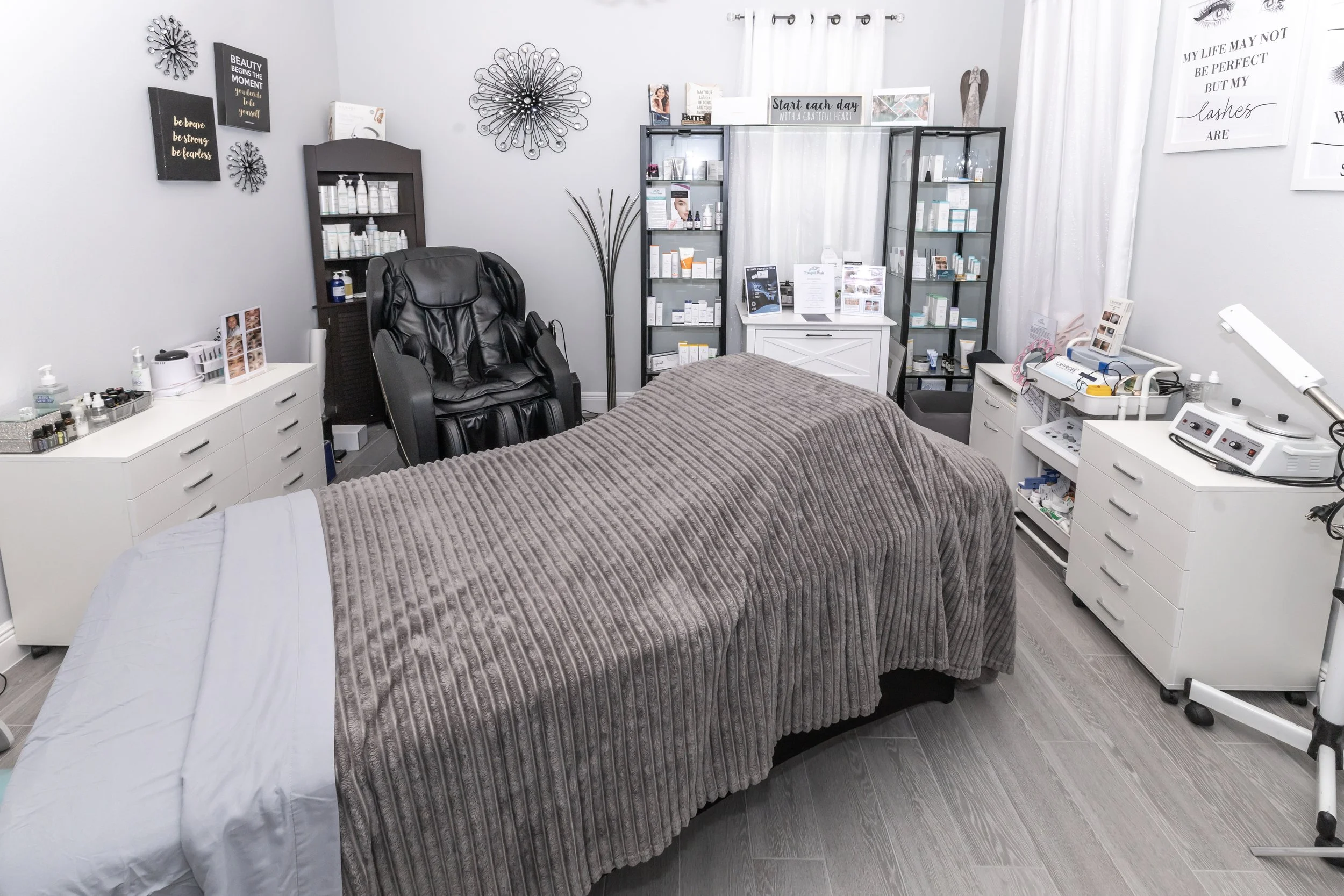 A medical or spa treatment room featuring a treatment bed with a gray blanket, surrounded by white cabinets, shelves with skincare products, a black massage chair, and various medical or skincare equipment.