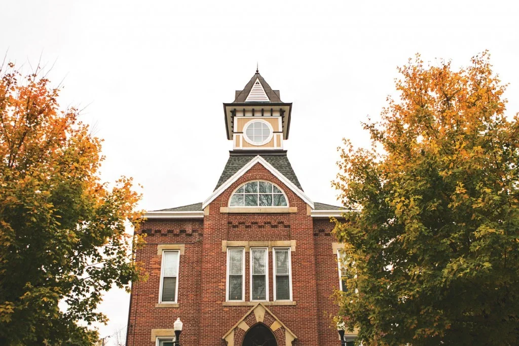 A historic red brick town hall building with a clock tower, surrounded by green trees, benches, and black lampposts with blue banners, under a partly cloudy sky.