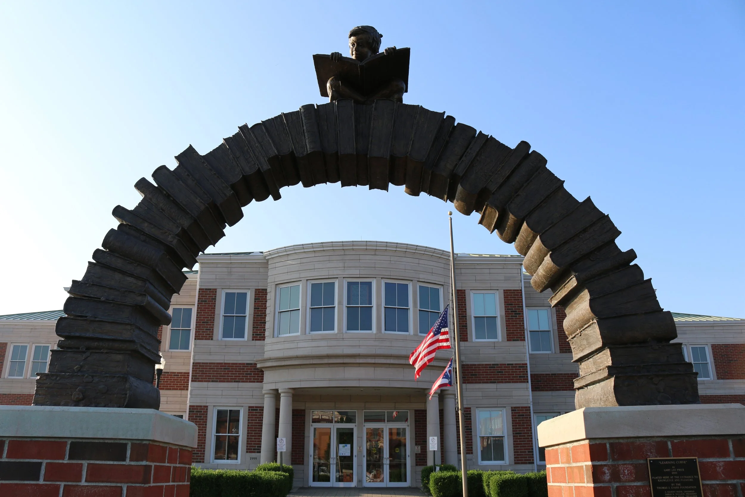 A Gothic-style stone arch with a statue on top, framing a building with a porch, American flag, and large windows.