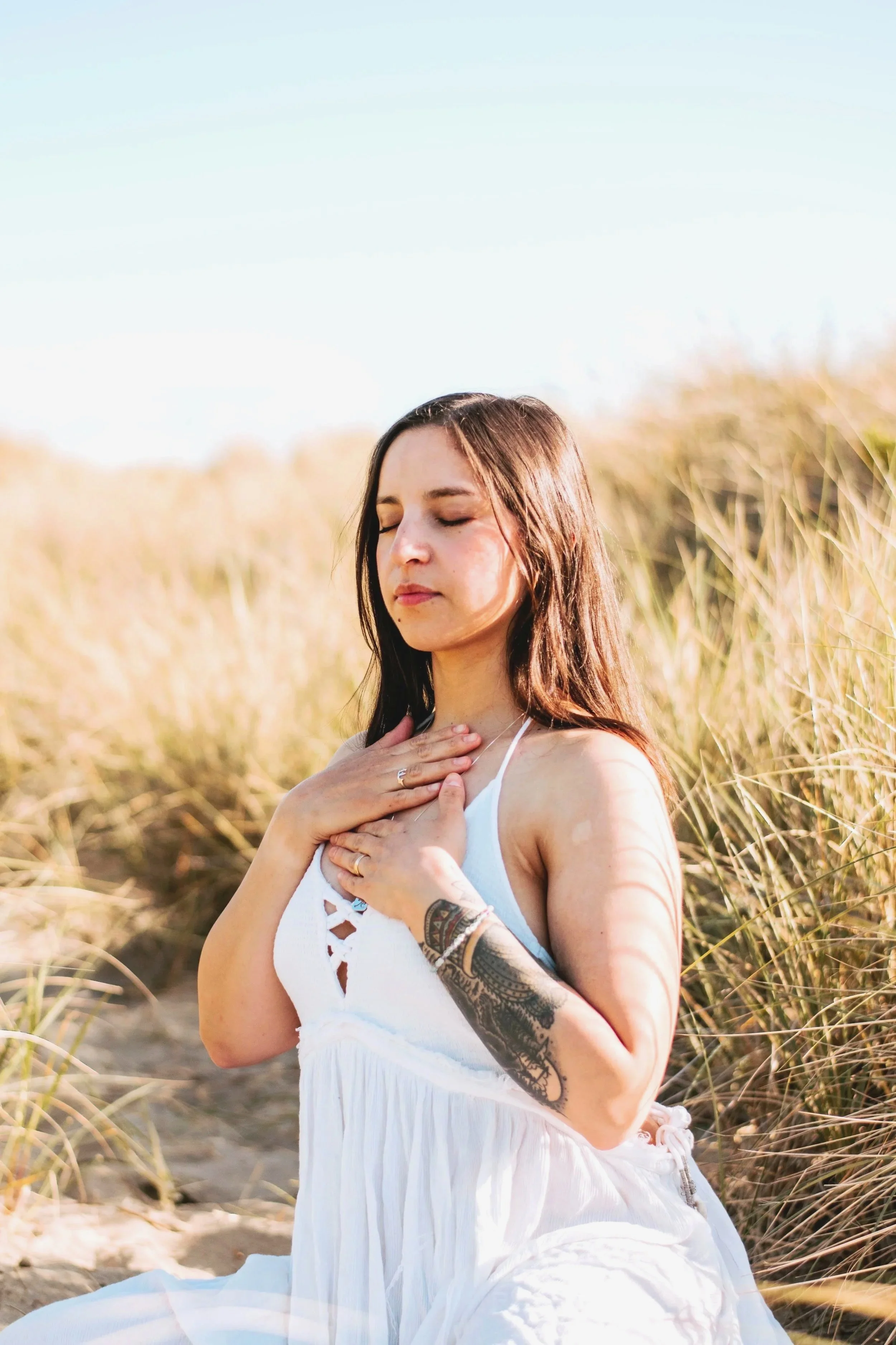 A woman with long brown hair and a tattoo on her arm, sitting outdoors in a field of tall grass, practicing meditation with her eyes closed and hands on her chest.