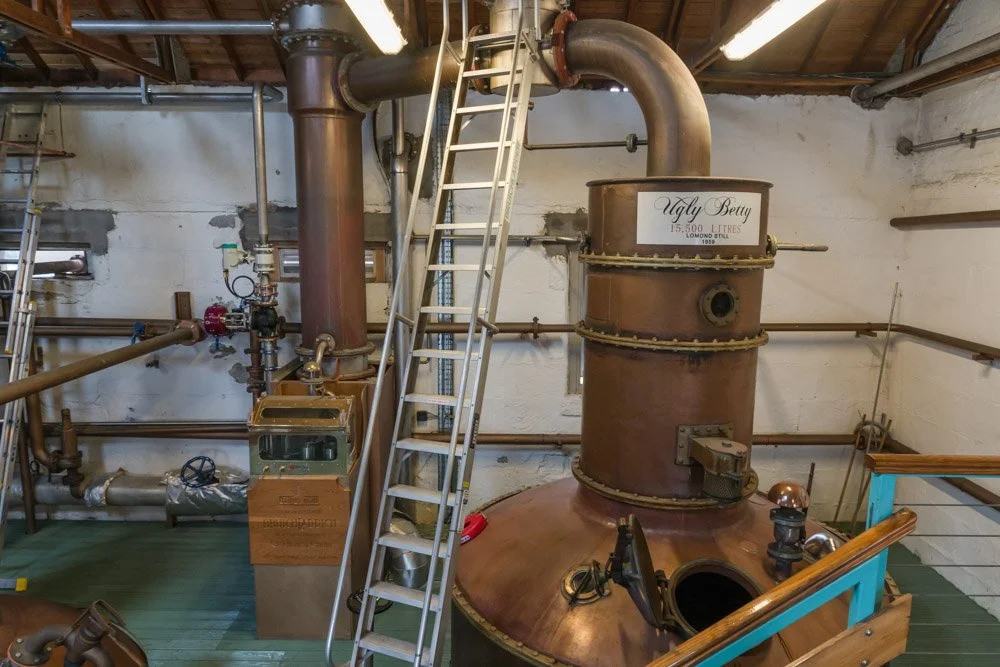 Industrial distillation apparatus with copper and metal piping, a large copper still labeled 'Ugly Betty' with a capacity of 15,500 liters, stairs, gauges, valves, and a control box in a workshop or distillery setting.