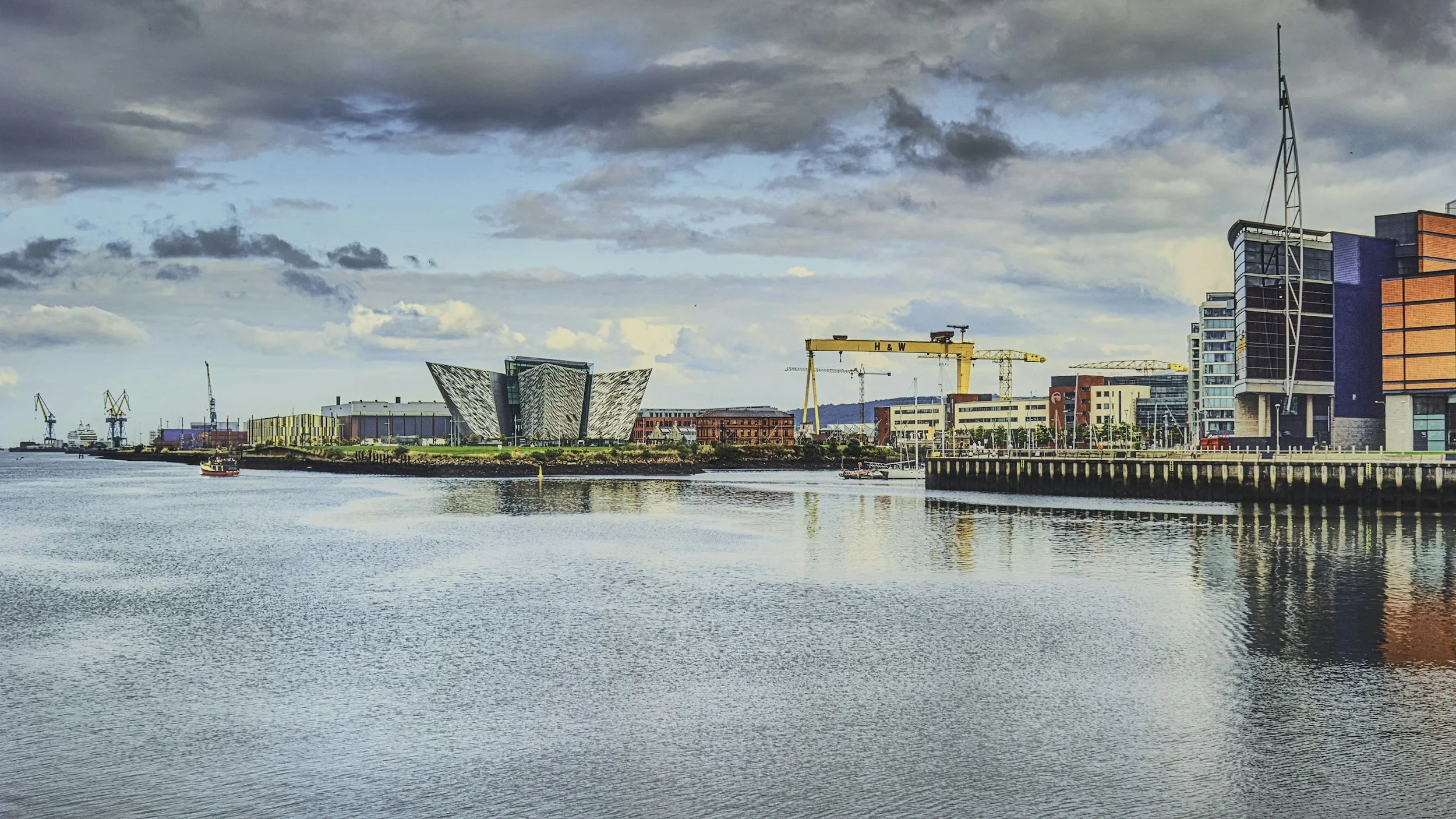A city skyline along a body of water with modern buildings, a crane, and a cloudy sky.