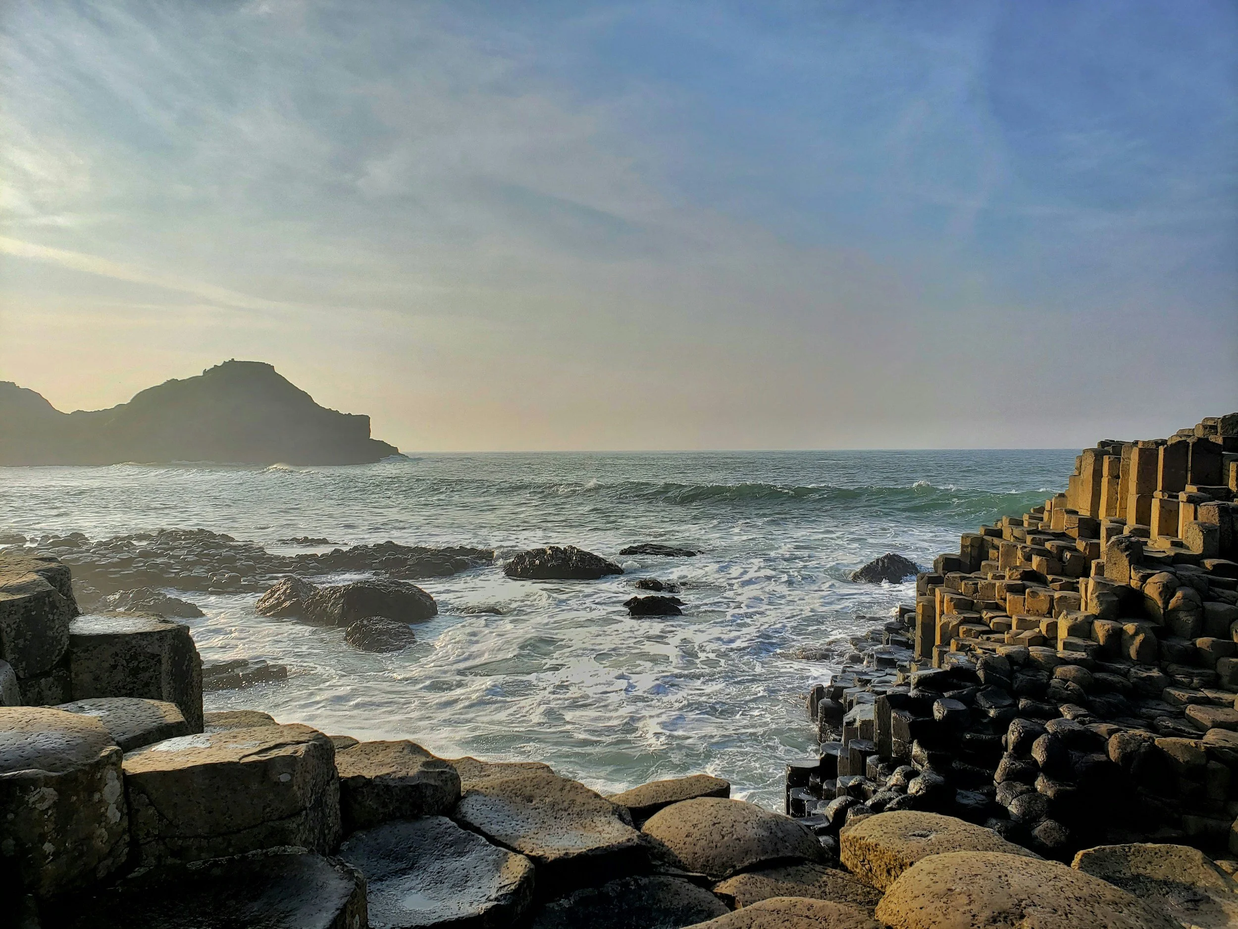 View of the ocean with waves crashing against rocks and basalt columns, with a hill or small mountain in the distance under a partly cloudy sky during sunset.