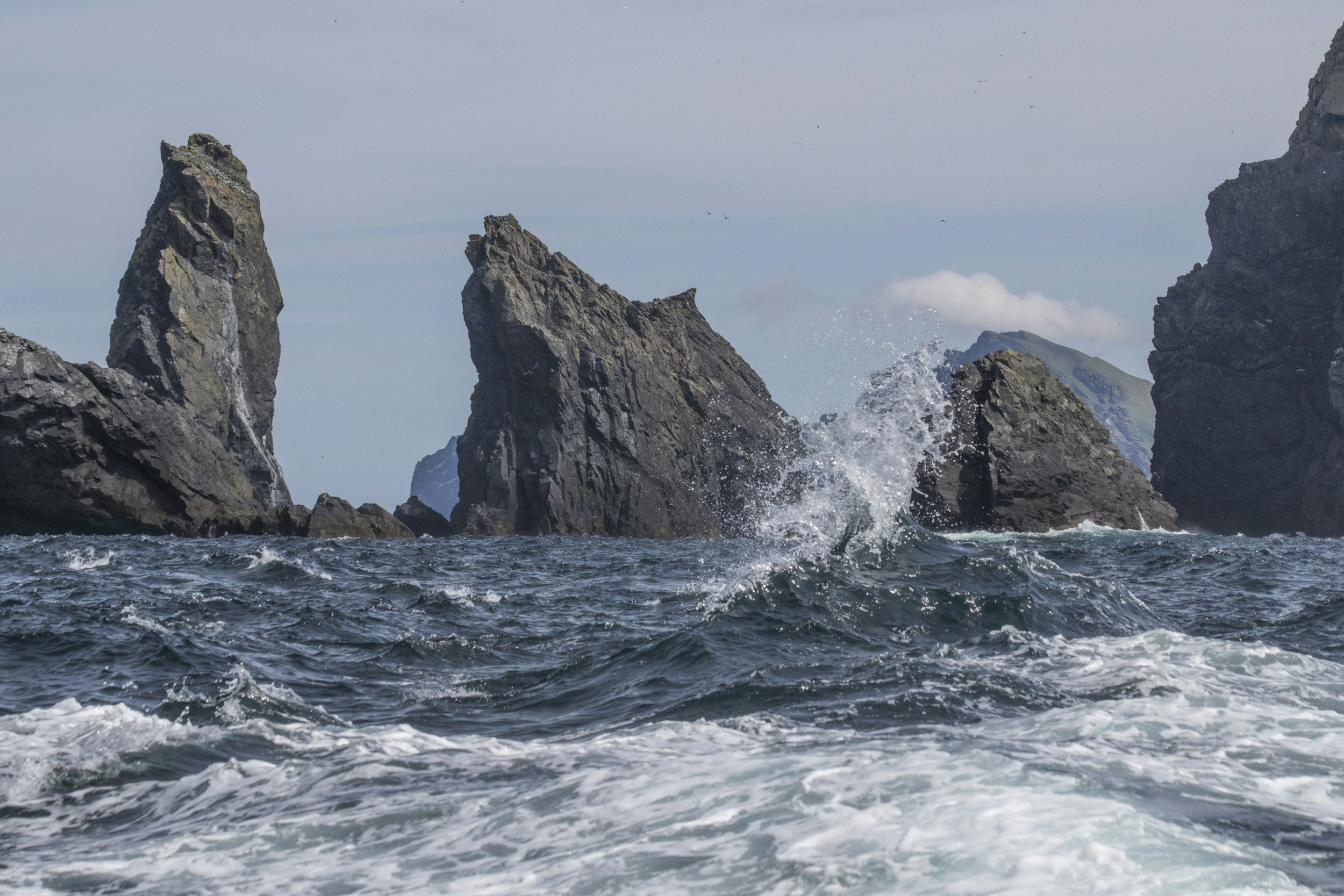Ocean waves crashing against large rocky sea stacks under a cloudy sky.