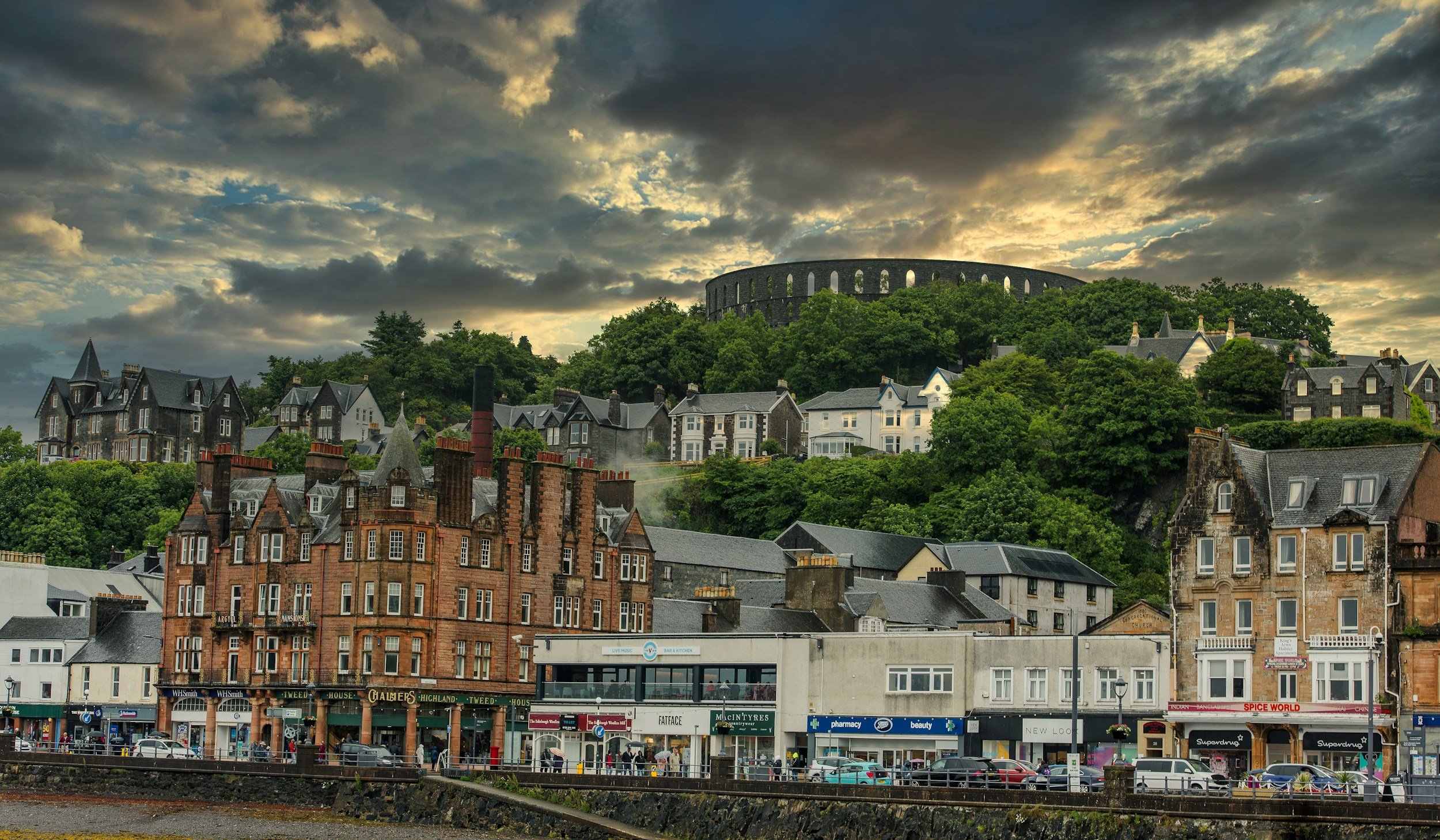 Cityscape with historic buildings at street level, commercial shops, and houses on a hill, with a fortress or large structure on a green hilltop in the background under a cloudy sky.