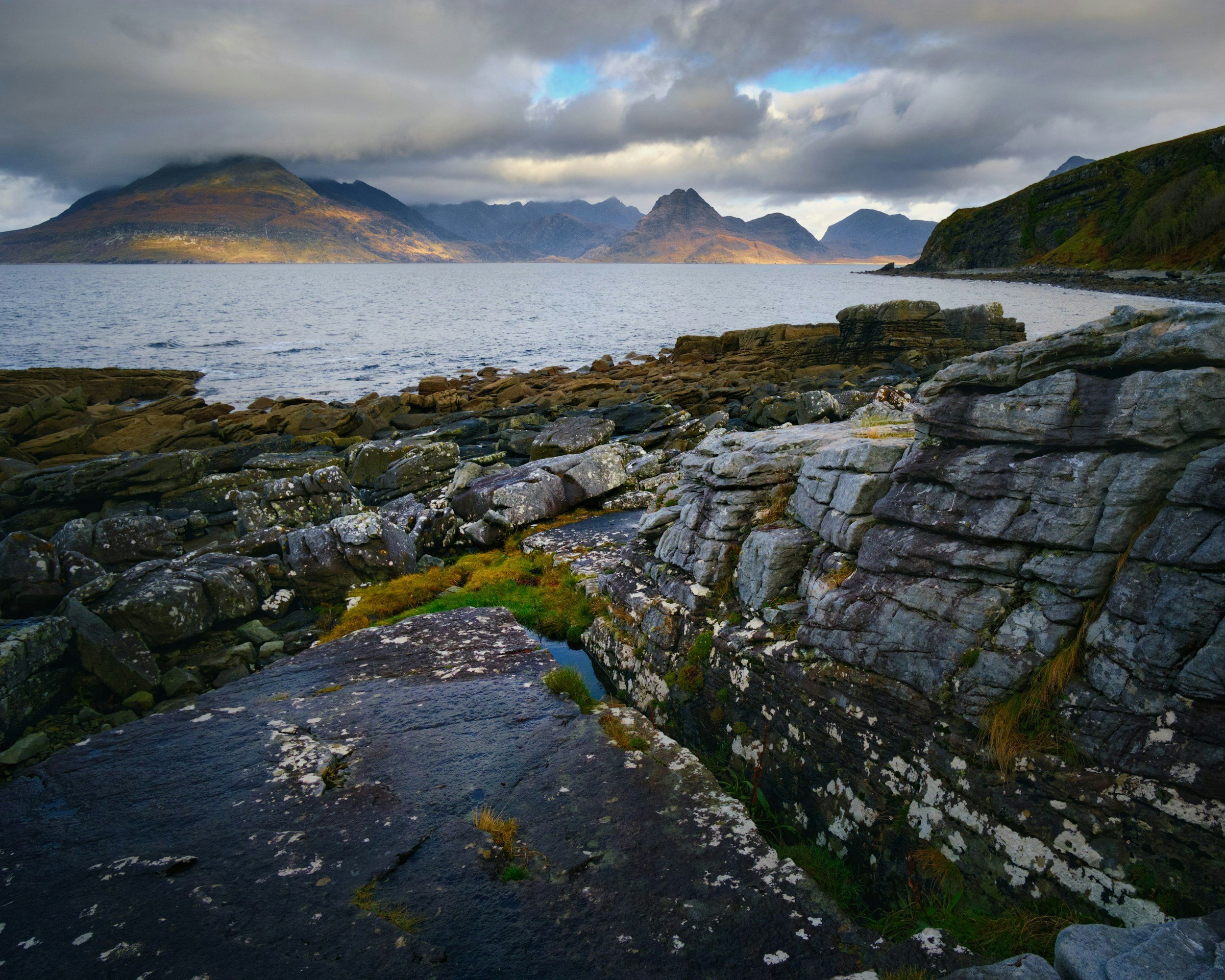 Rocky shoreline with large gray rocks and patches of green moss, overlooking a body of water with mountains in the background and a partly cloudy sky.
