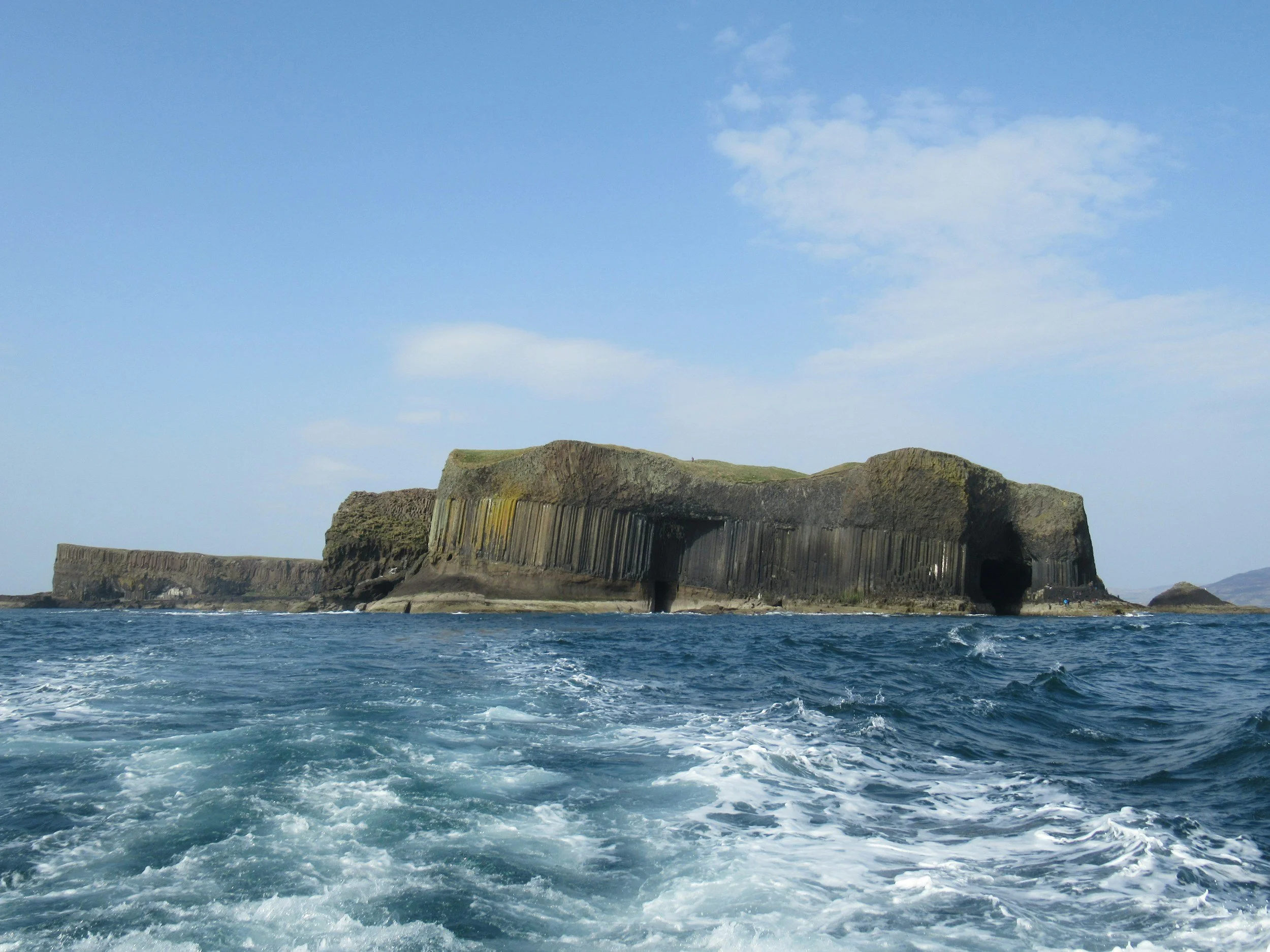 Ocean view showing a large rocky island with caves and cliffs, under a blue sky with some clouds.