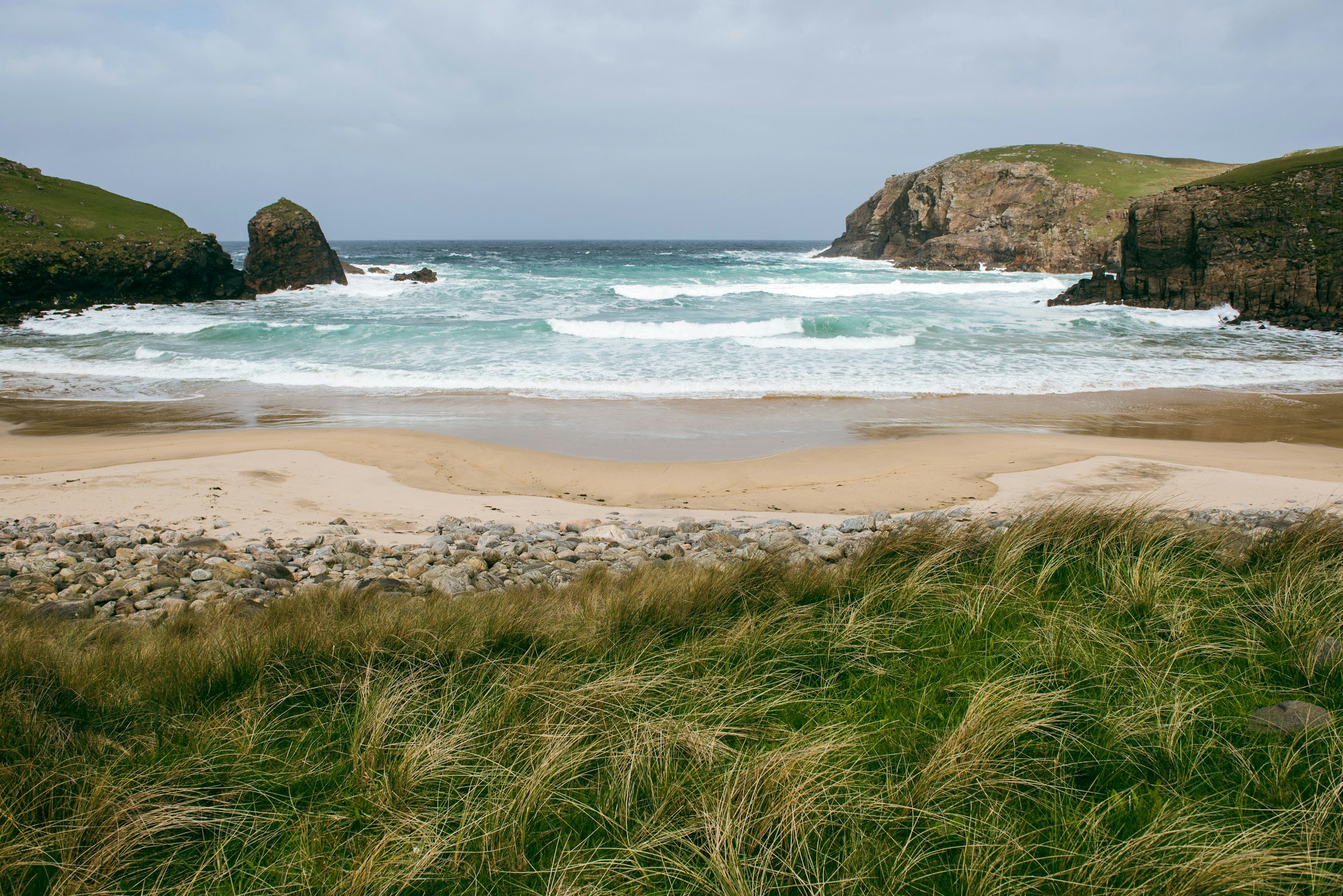 Ocean waves crashing against large rocky sea stacks under a cloudy sky.