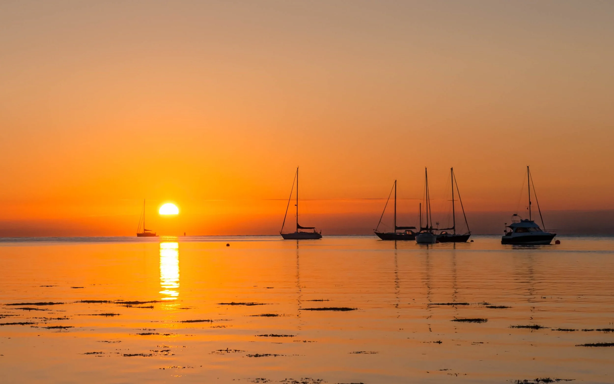 Sunset over calm sea with five sailboats anchored, warm orange and pink sky reflected on water, distant horizon.