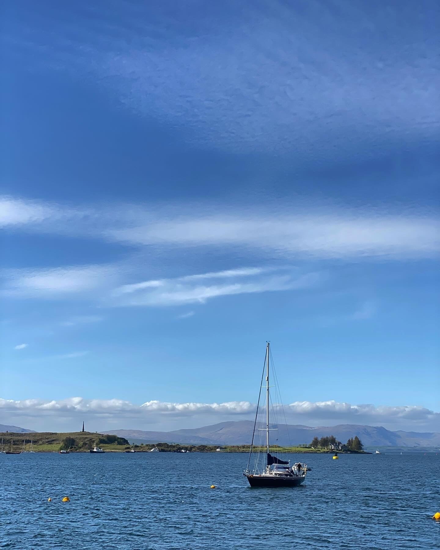 A sailboat floating on a calm body of water with a scenic landscape in the background, including hills, mountains, and a partly cloudy sky.