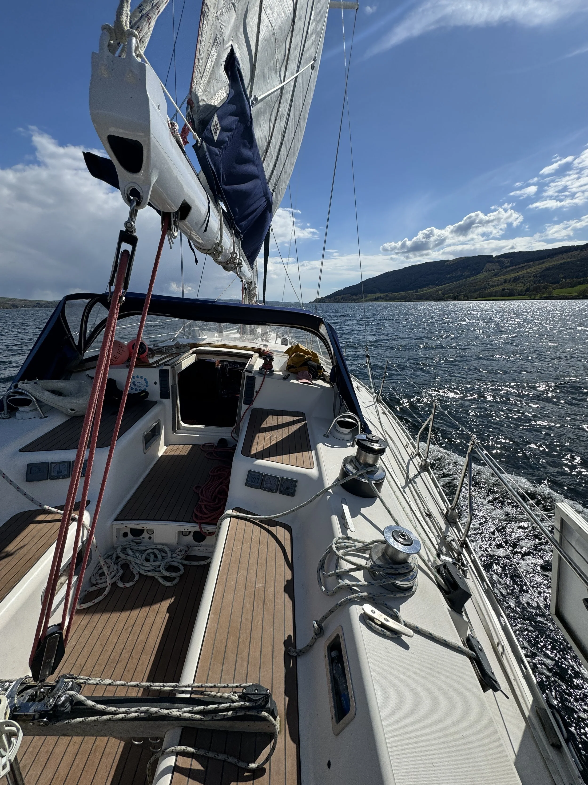 View of a sailboat sailing on a lake with green hills in the background on a sunny day with blue sky and some clouds.