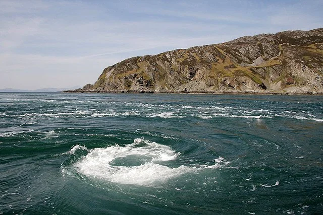 Ocean with a whirlpool near the shoreline and hills in the background.
