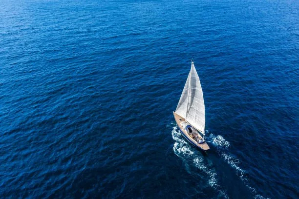 A sailboat with white sails sailing on the open blue ocean from an aerial perspective.