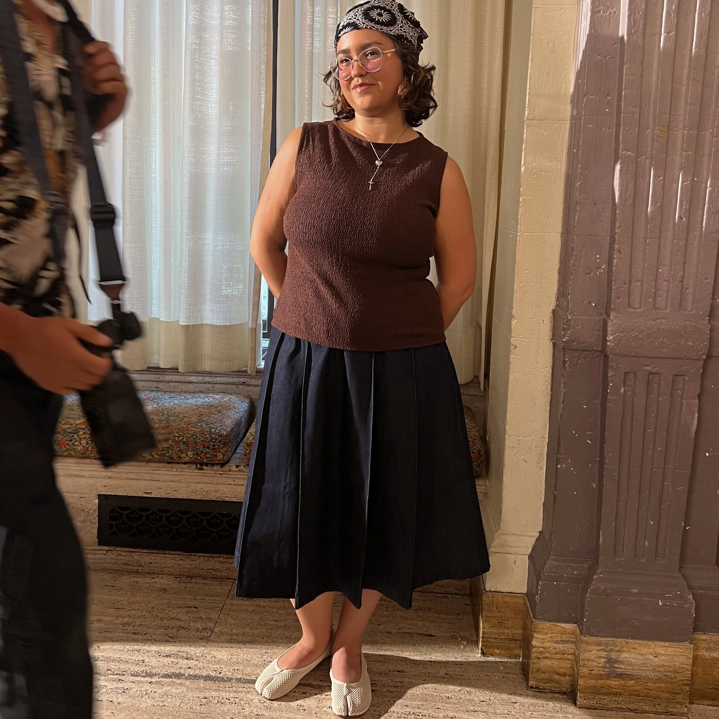 A woman standing inside near a curtain with a smile, wearing glasses, a patterned bandana, a sleeveless brown top, a necklace, a black skirt, and white shoes, with her hands behind her back.